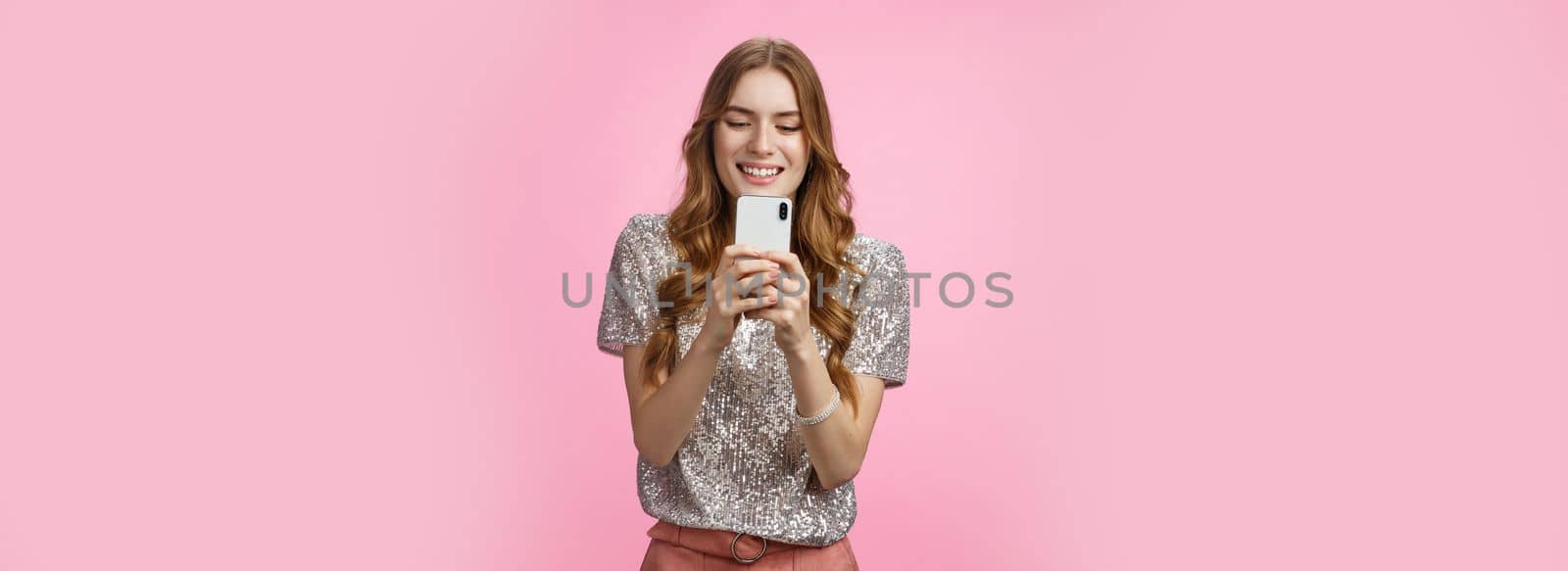 Charming glamour sociable stylish caucasian female using smartphone looking mobile phone screen look gadget display amused writing post upload photo social web page texting, pink background.