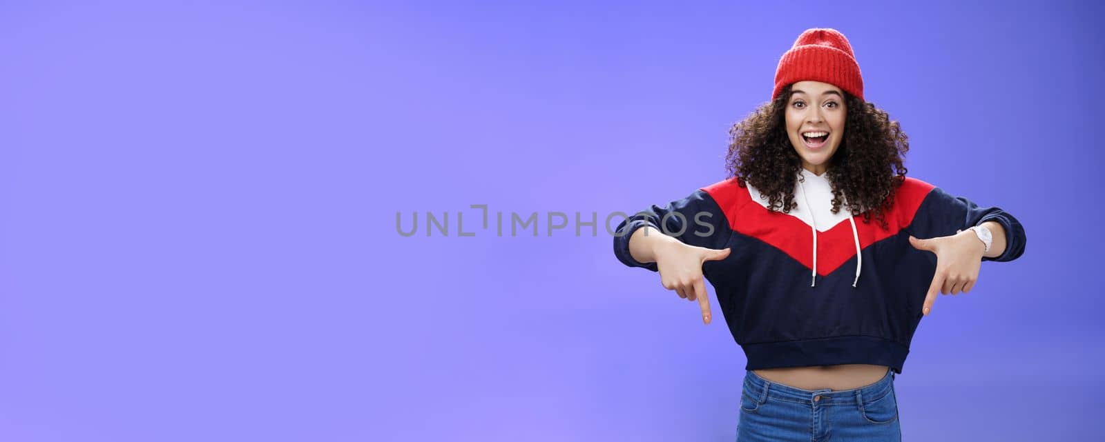 Hurry up and chek it out. Portrait of charismatic joyful and excited young cute curly-haired girl in beanie and stylish outfit pointing down, smiling broadly inviting join fun over blue background.