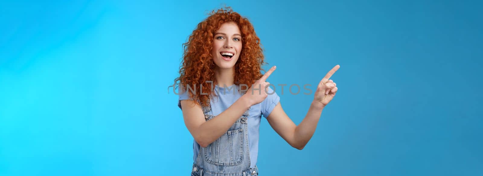 Hey check this out. Enthusiastic redhead curly stylish summer girl inviting beach party pointing upper left corner directing awesome location smiling broadly discuss awesome promo blue background by Benzoix