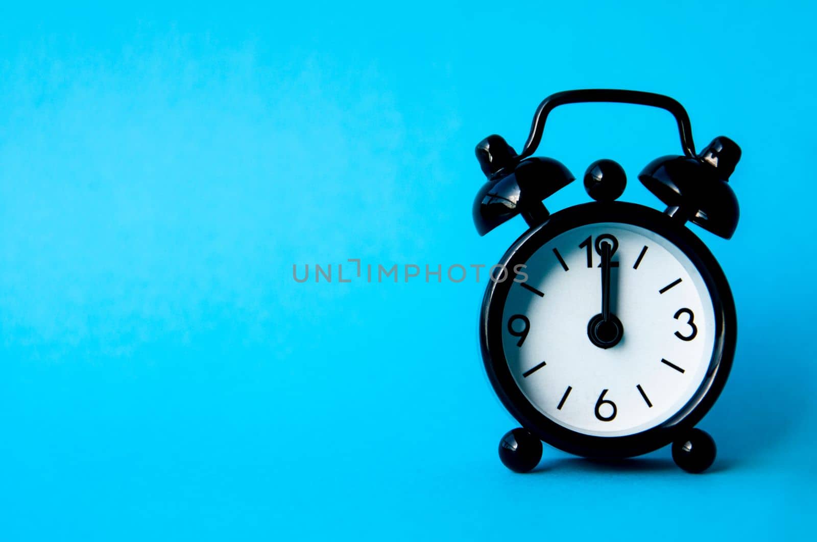 Black alarm clock isolated on blue background. The clock set at 12pm. Copy space.
