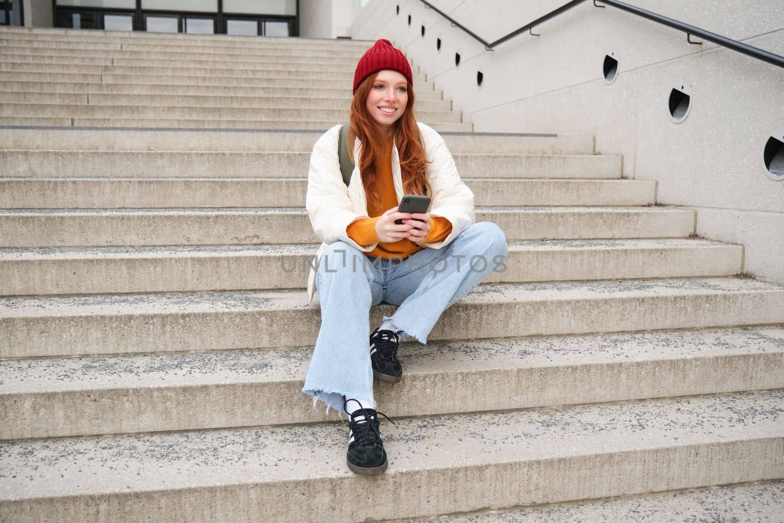 City lifestyle and mobile phone. Smiling college girl with red hair, sits on public stairs on street with smartphone, reads message, uses social media app.