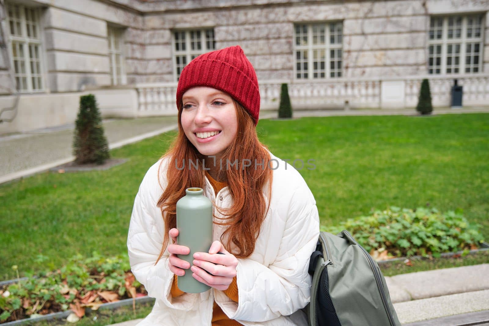 Smiling redhead girl rests in park, sits on bench with backpack, drinks from thermos, enjoys hot drink from flask and looks relaxed.