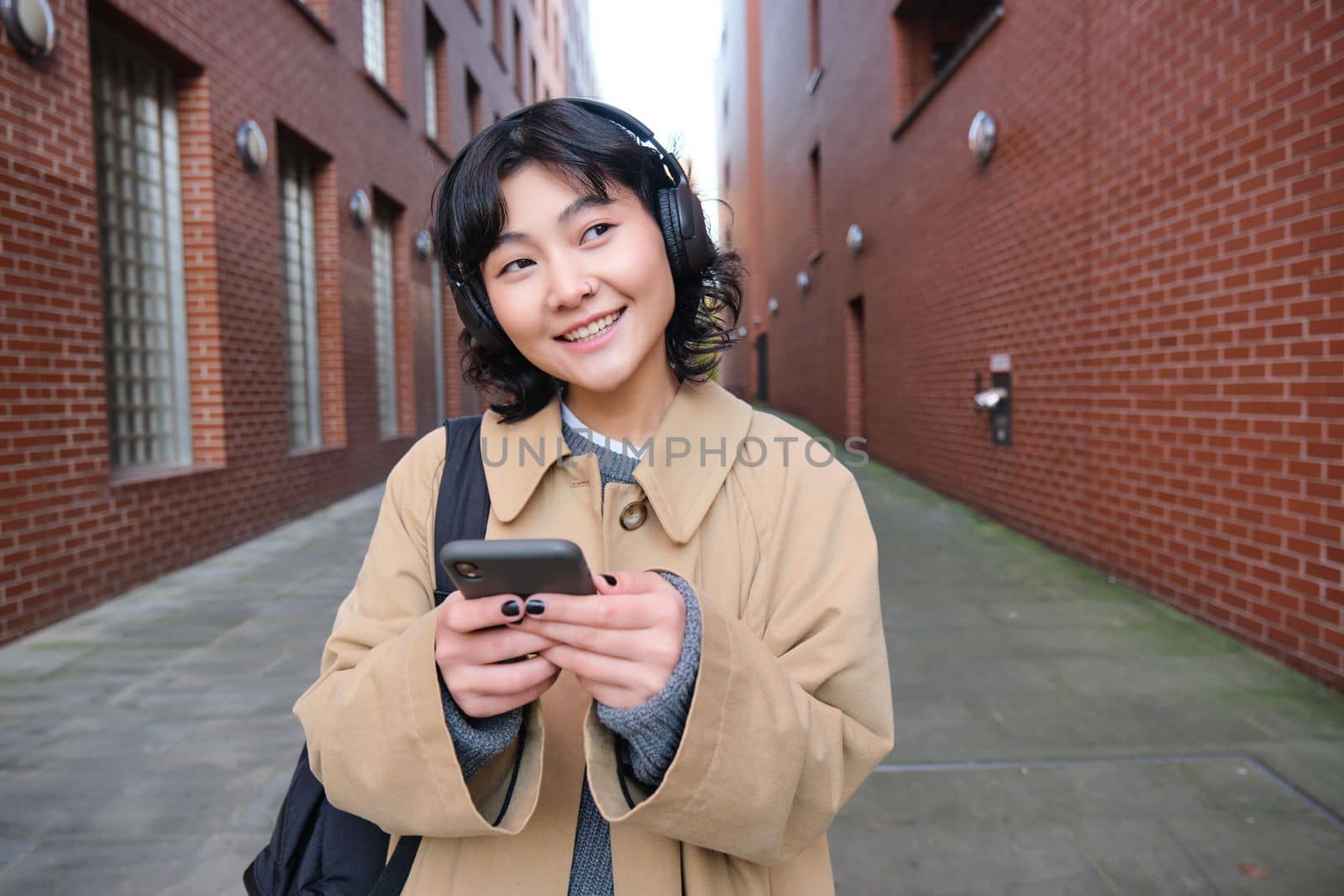 Portrait of young korean woman walking around city with backpack and headphones, listens music, looks at smartphone, uses phone application on streets.