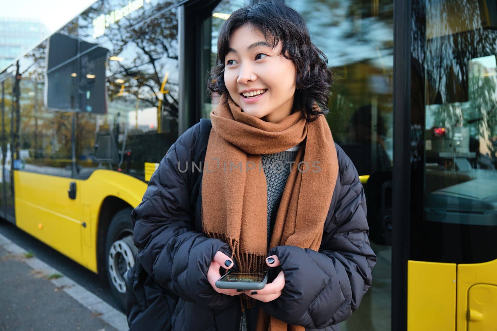 Young beautiful woman standing on bus stop, texting message on smartphone, holding mobile phone, checking her schedule, buying ticket online, wearing winter clothes by Benzoix