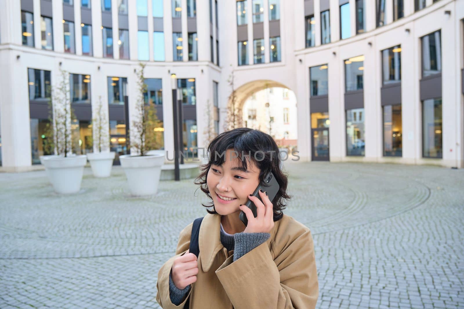 Cellular technology. Young korean woman talks on mobile phone, makes a phone call on her way home, walks down street, city centre, has telephone conversation.