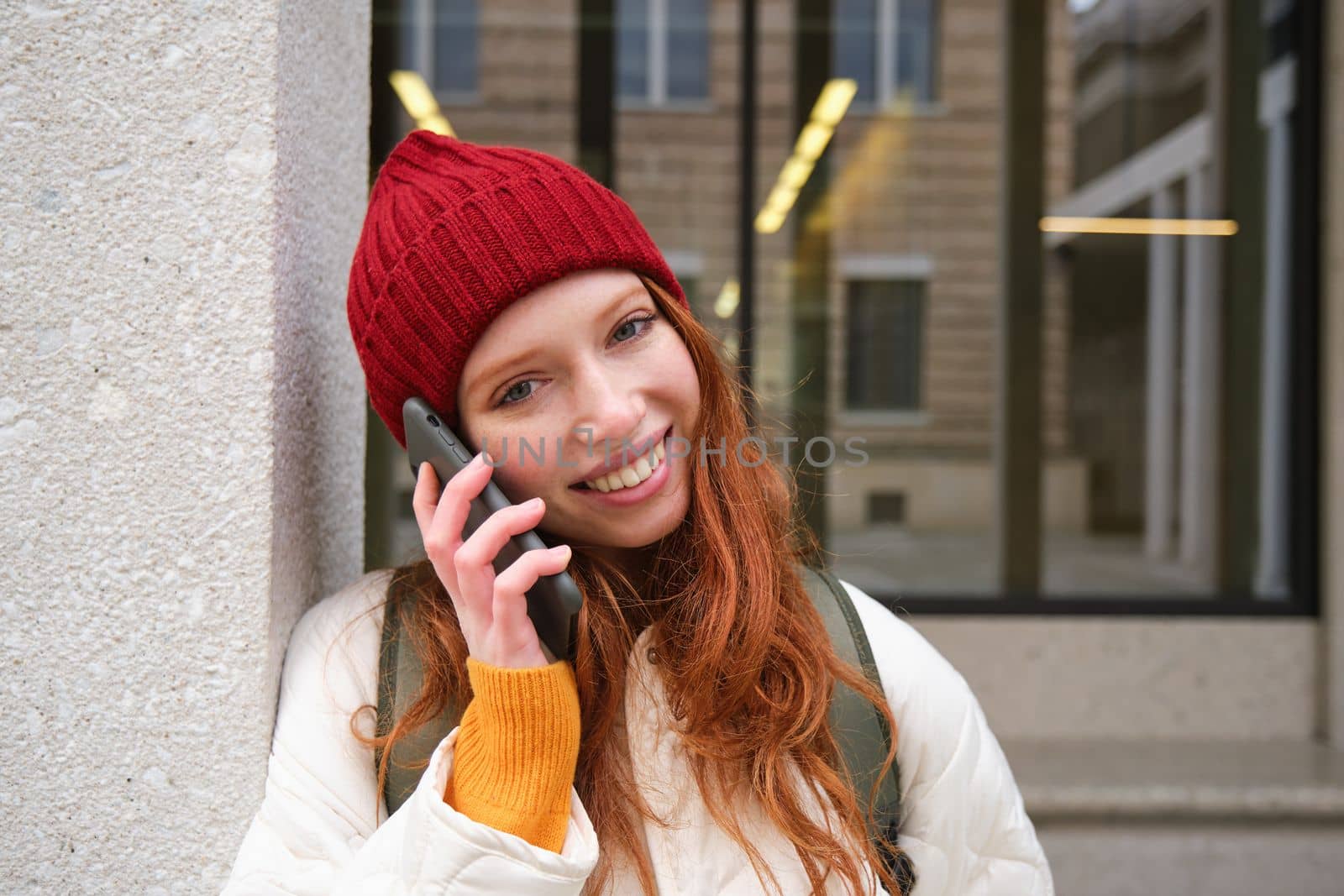 Happy beautiful girl, redhead tourist talks on mobile phone, making a call abroad, has conversation on smartphone, standing with backpack outdoors.