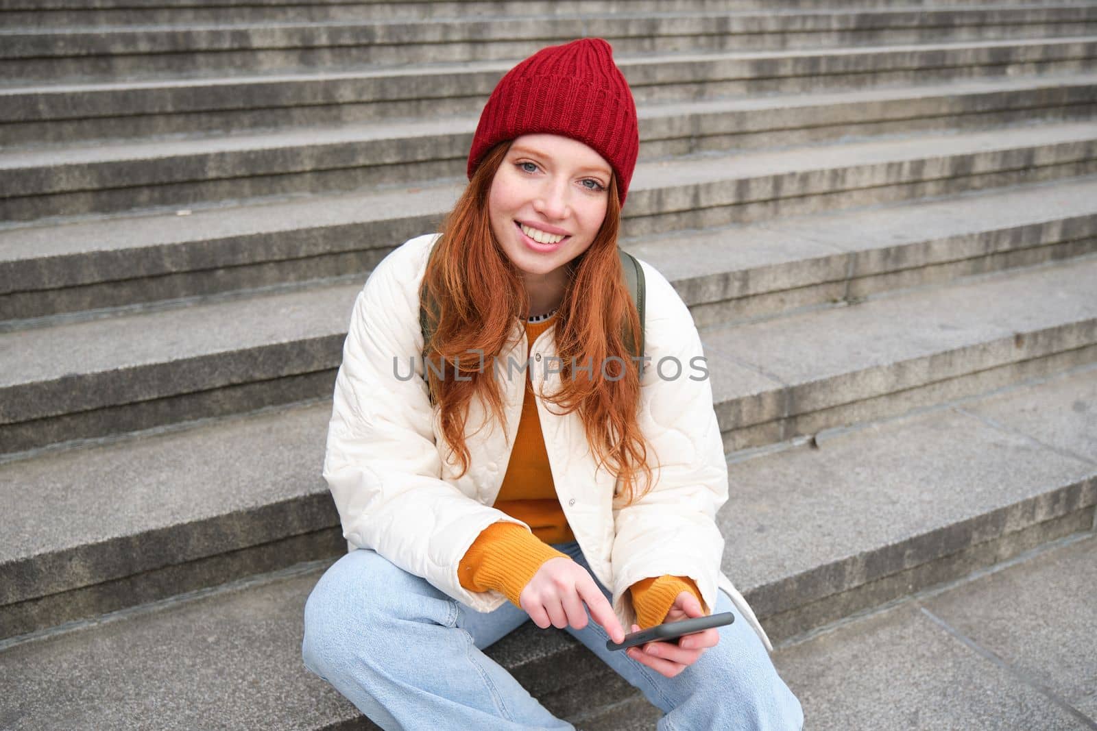Stylish young redhead woman, talking on mobile phone app, using social media application, looking for something online on smartphone, sits on stairs outdoors by Benzoix