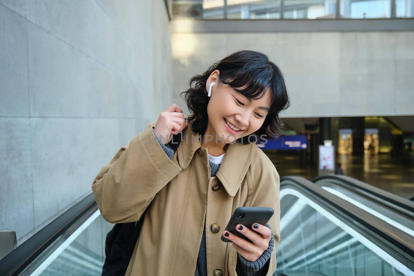 Portrait of cute korean girl in trench coat, going up escalator, holds mobile phone and smiles pleased, commutes, watches video or listens music with wireless headphones.
