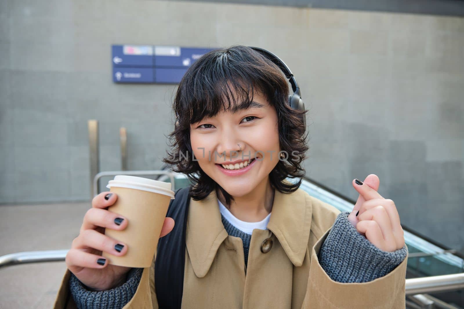 Portrait of stylish asian woman in headphones, drinks coffee to go and smiles, enjoys cappuccino while commutes, stands on street by Benzoix