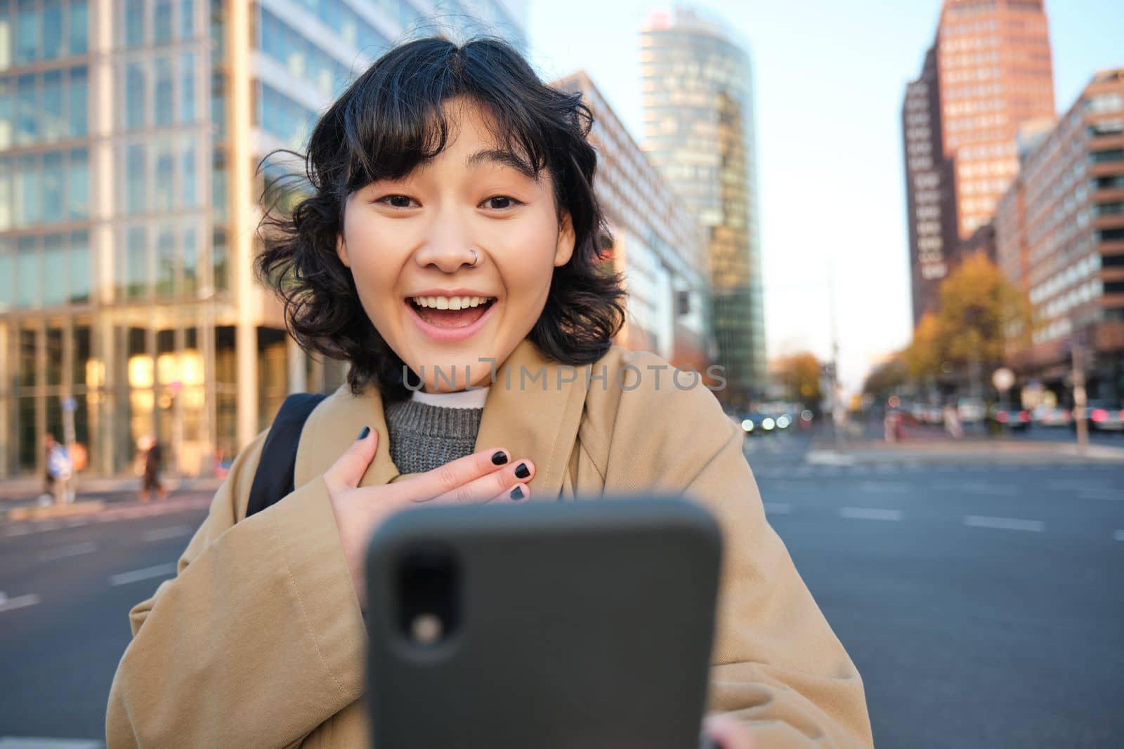 Portrait of asian girl looks surprised at smartphone screen, video chats and hears amazing news, looks impressed, stands on sunny street of city centre.