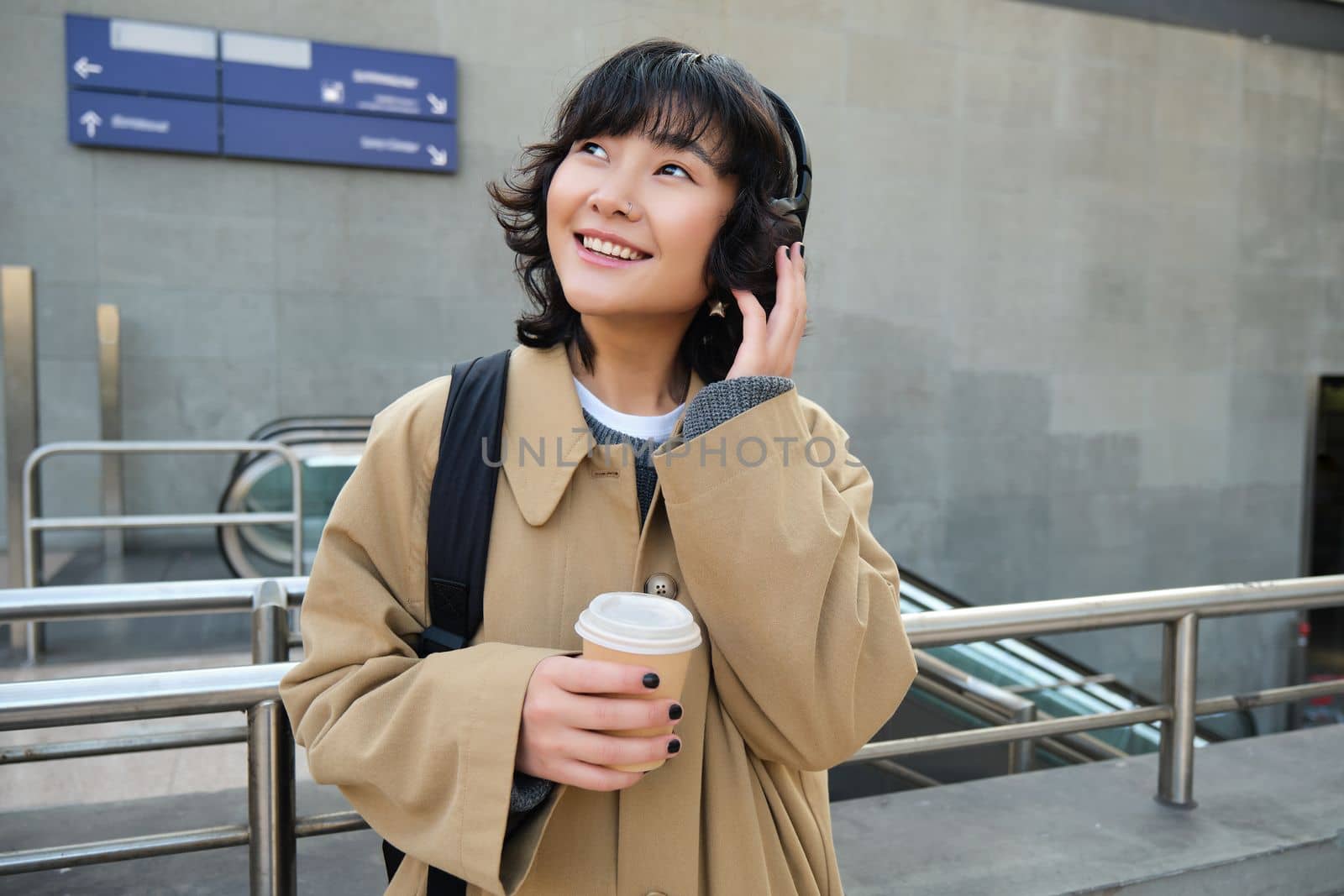 Portrait of young student, girl in headphones, drinks coffee, stands on street with backpack, commutes to university or college, smiles happily.