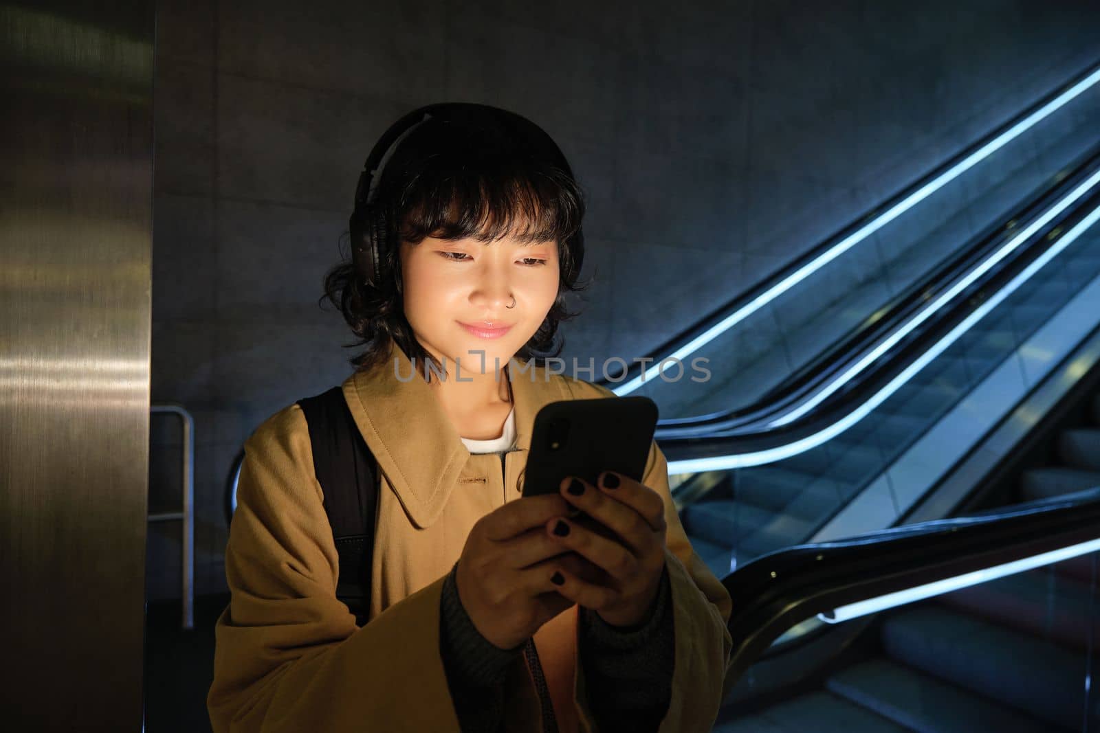 Portrait of young smiling asian girl, stands near escalator and waits for someone, reads message, checks map on smartphone, listens music in headphones.