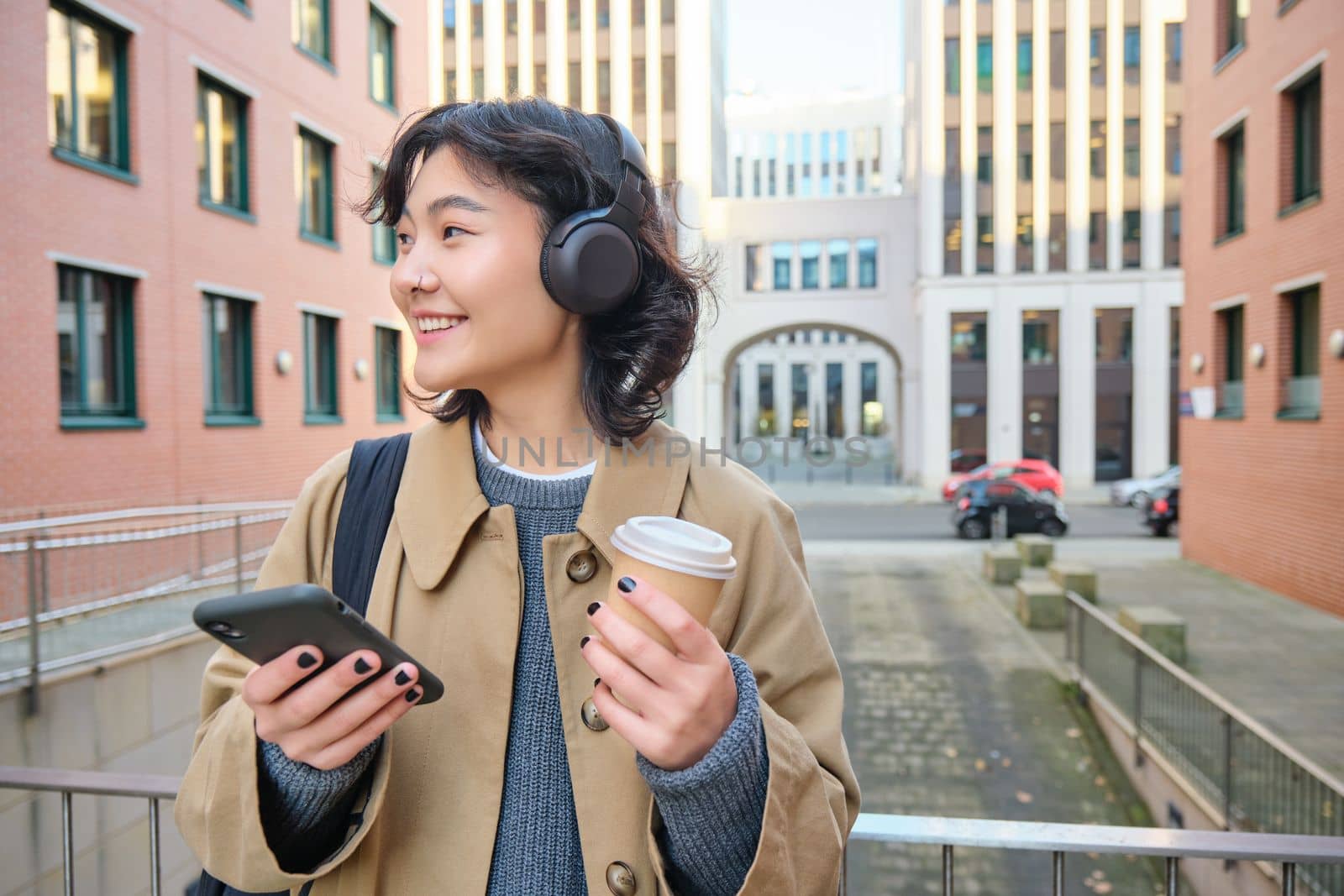 Young woman, tourist in headphones looks at smartphone, drinks coffee to go, checks mobile phone app, listens music and travels. Copy space
