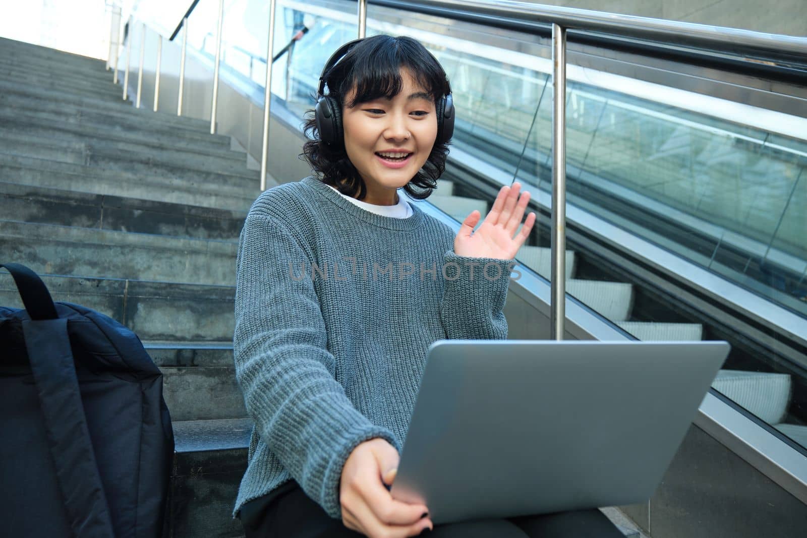 Smiling young student, wears headphones, sits on street stairs and waves hand at her laptop, connects to video chat, conference or training session on remote.