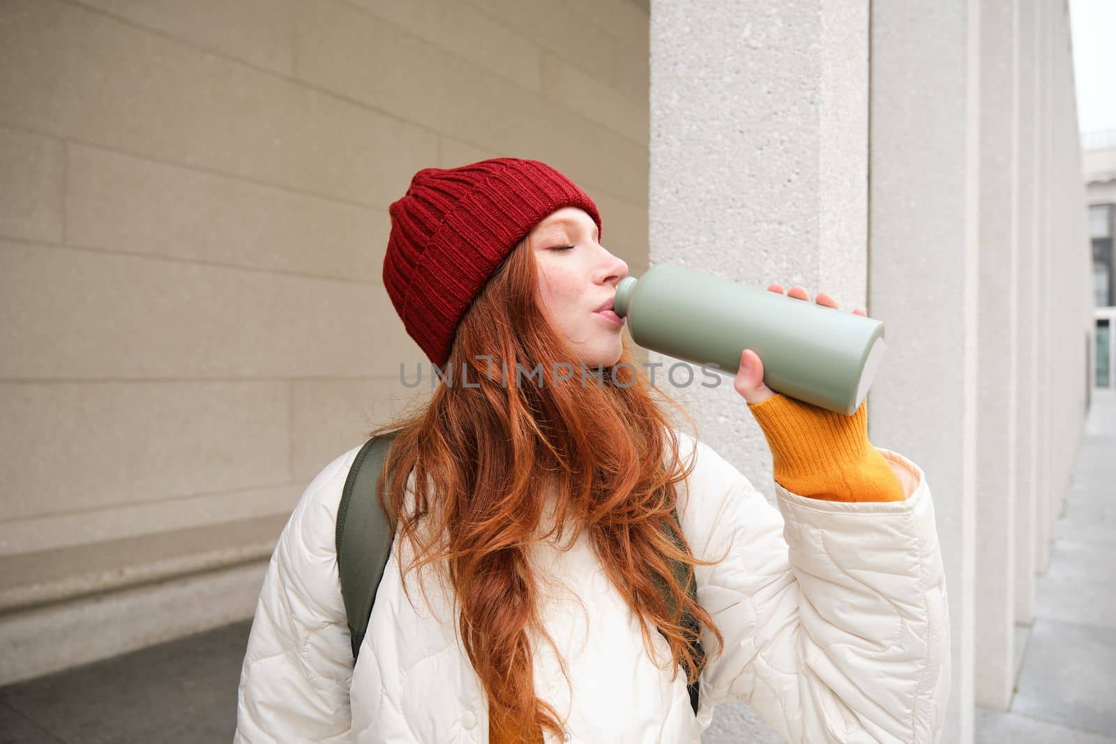 Beautiful redhead woman drinking hot tea or coffee from thermos, female tourist enjoys warm drink, rests during her journey around town by Benzoix