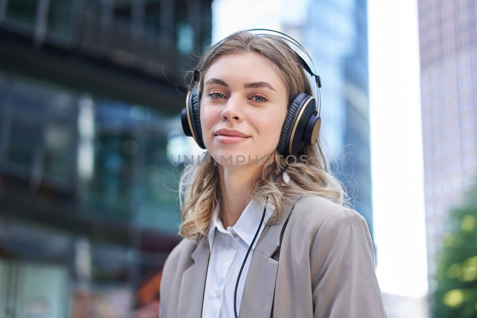 Portrait of smiling corporate woman, girl in suit listening music in headphones, sitting in city centre by Benzoix