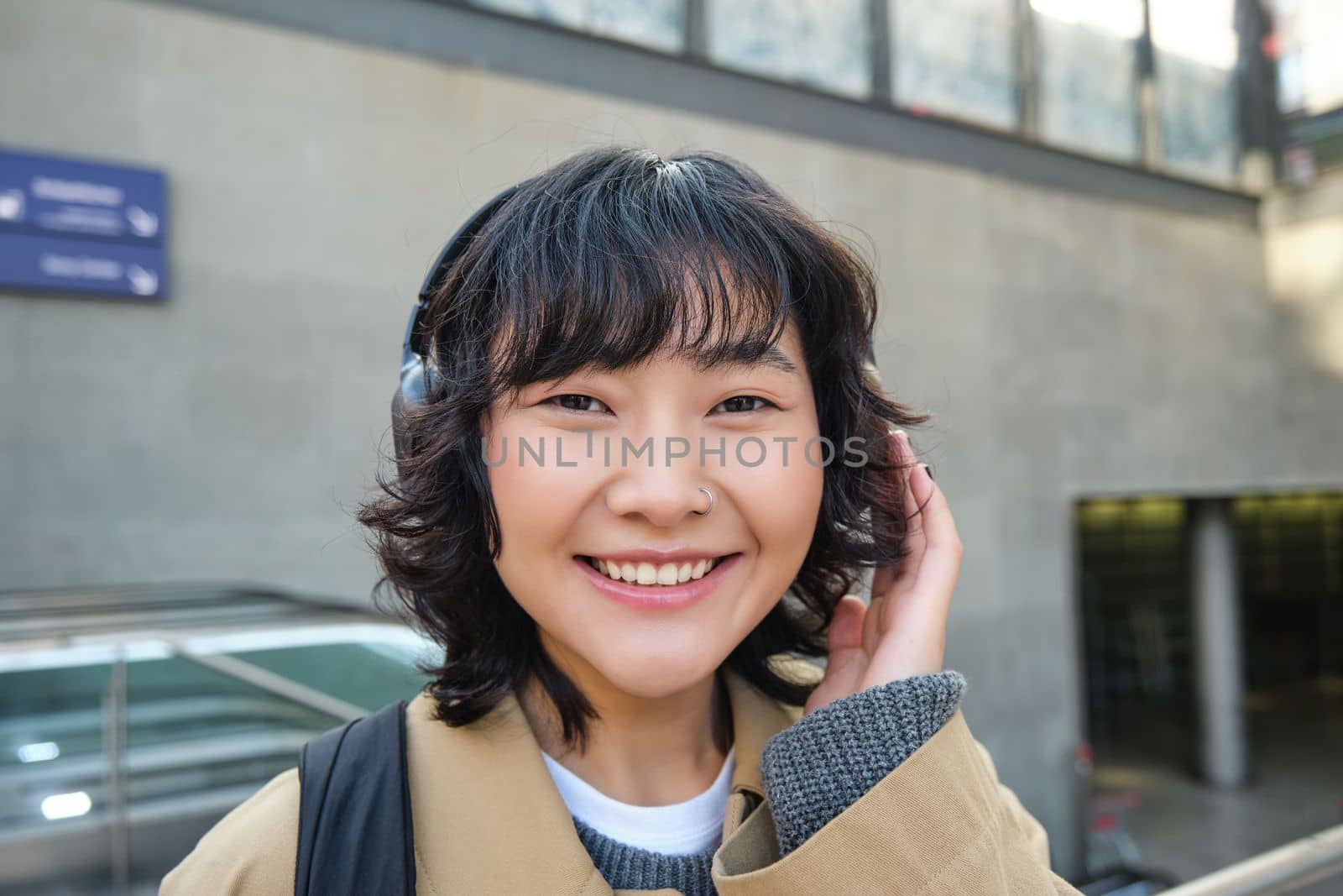Close up of smiling brunette girl in headphones, listens music, travels around city, commutes to work, stands on a street by Benzoix