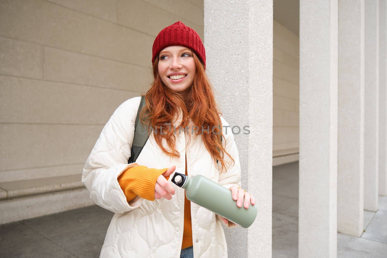 Young happy redhead woman in red hat, drinking from thermos, warming up with hot drink in her flask while walking around city, tourist relaxes with warm refreshemtn.