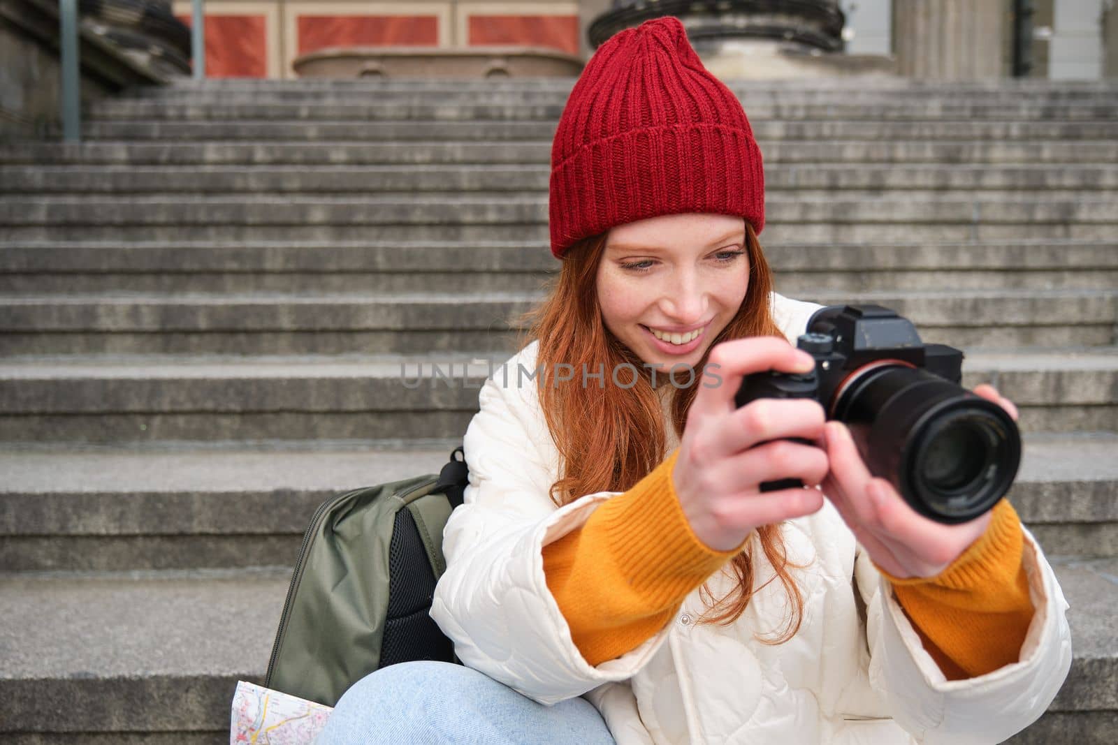 Portrait of young photographer girl, sits on stairs with professional camera, takes photos outdoors, making lifestyle shooting.