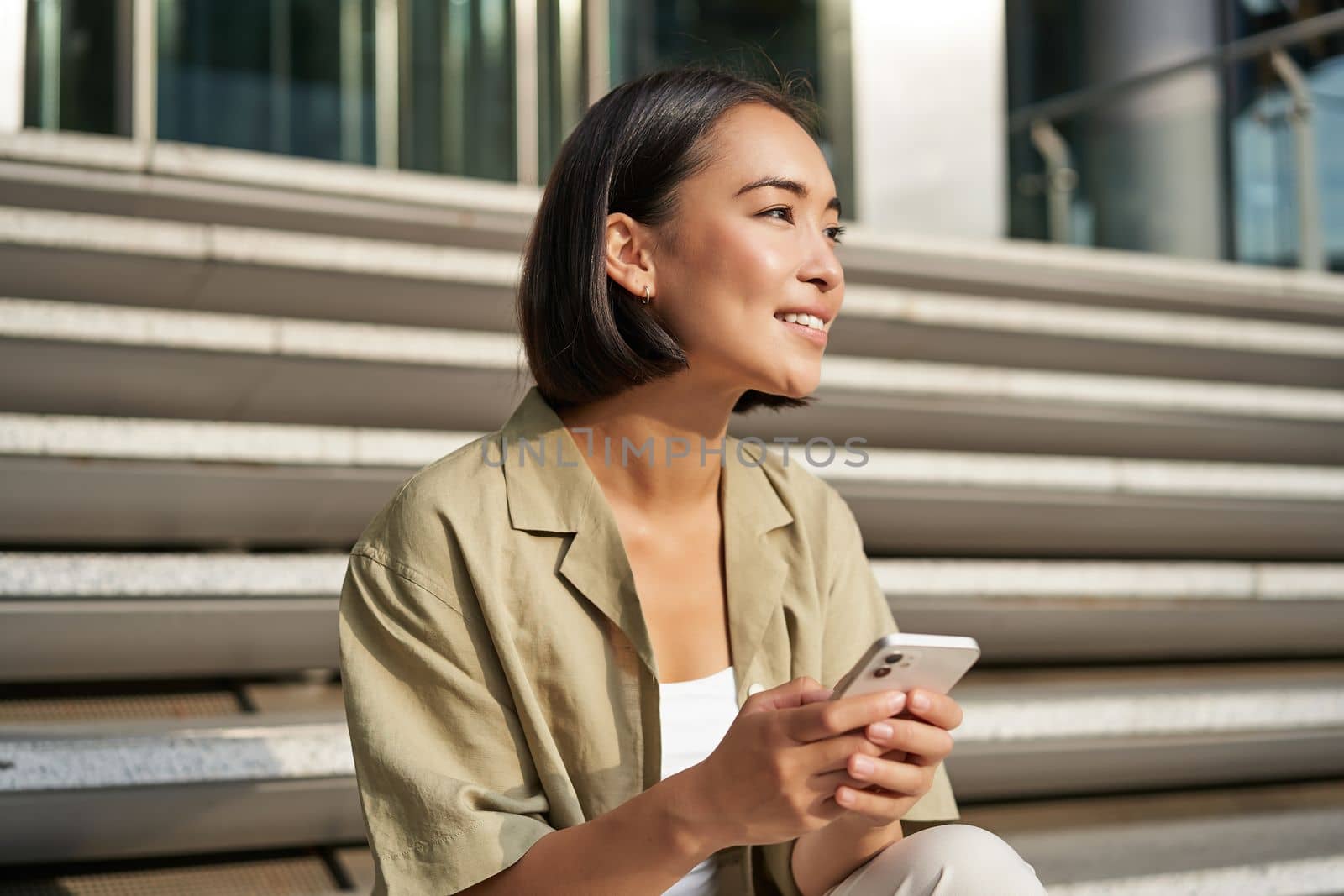 People and technology. Smiling beautiful asian woman sitting on stairs in city, holding mobile phone. Girl with smartphone rests outside by Benzoix