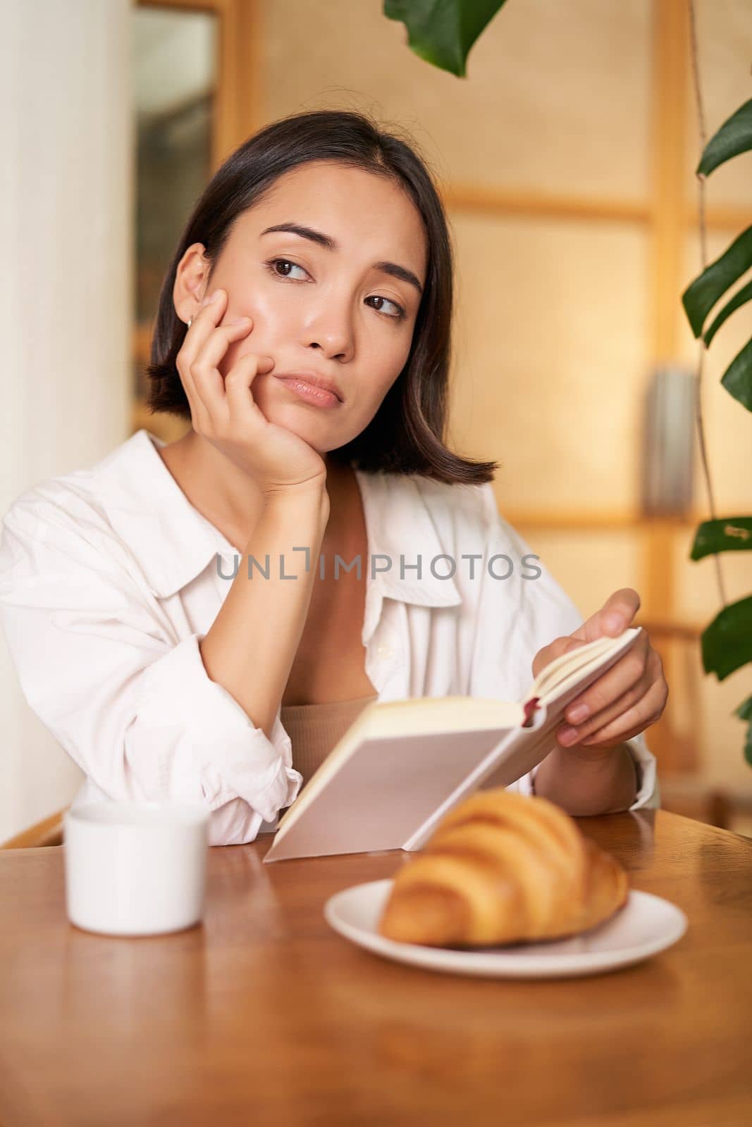 Vertical shot of asian girl sits alone in cafe, reads book and looks upset, drinks coffee with croissant.