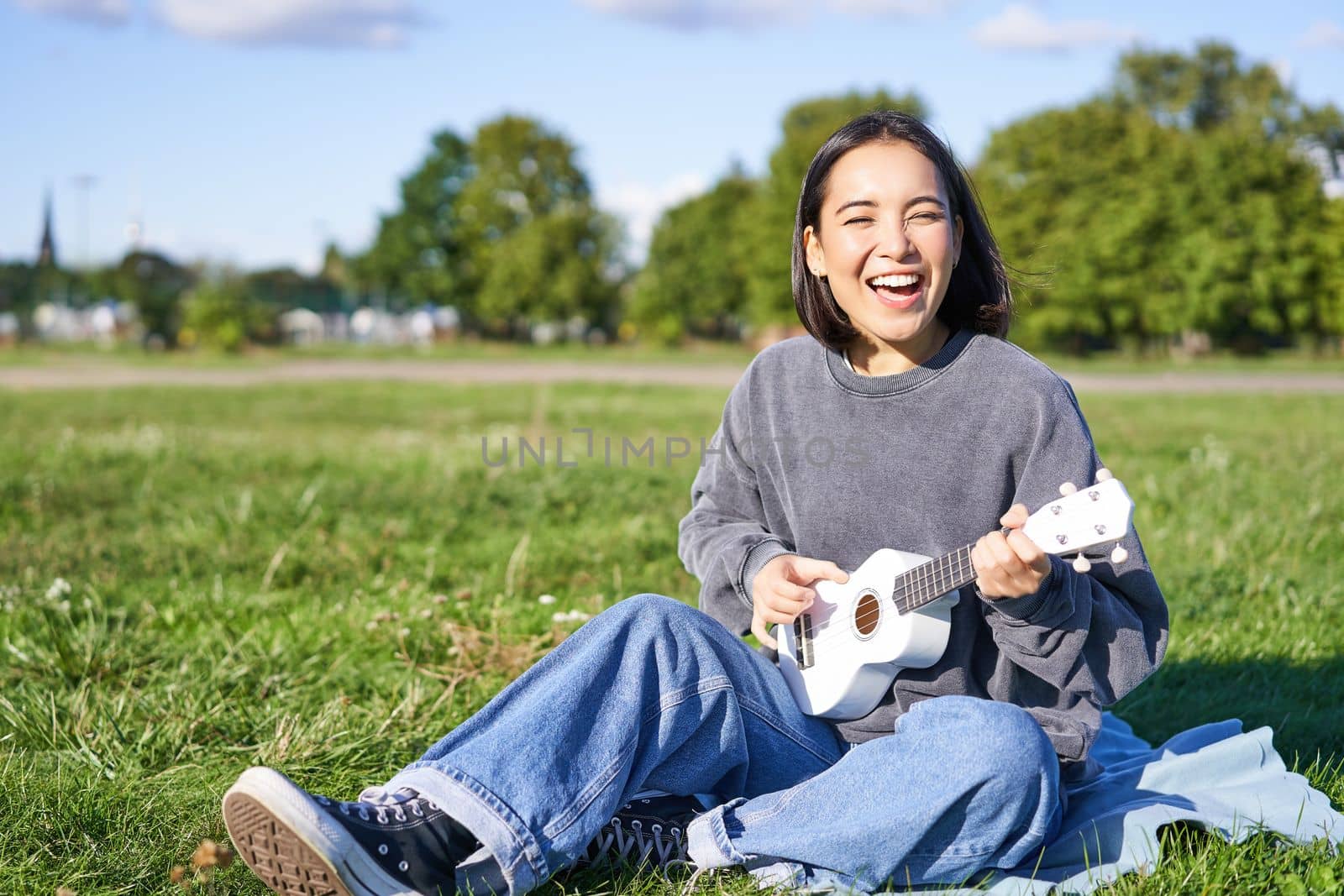 Portrait of beautiful asian woman singing, playing ukulele guitar in park, sitting alone on grass on sunny day.