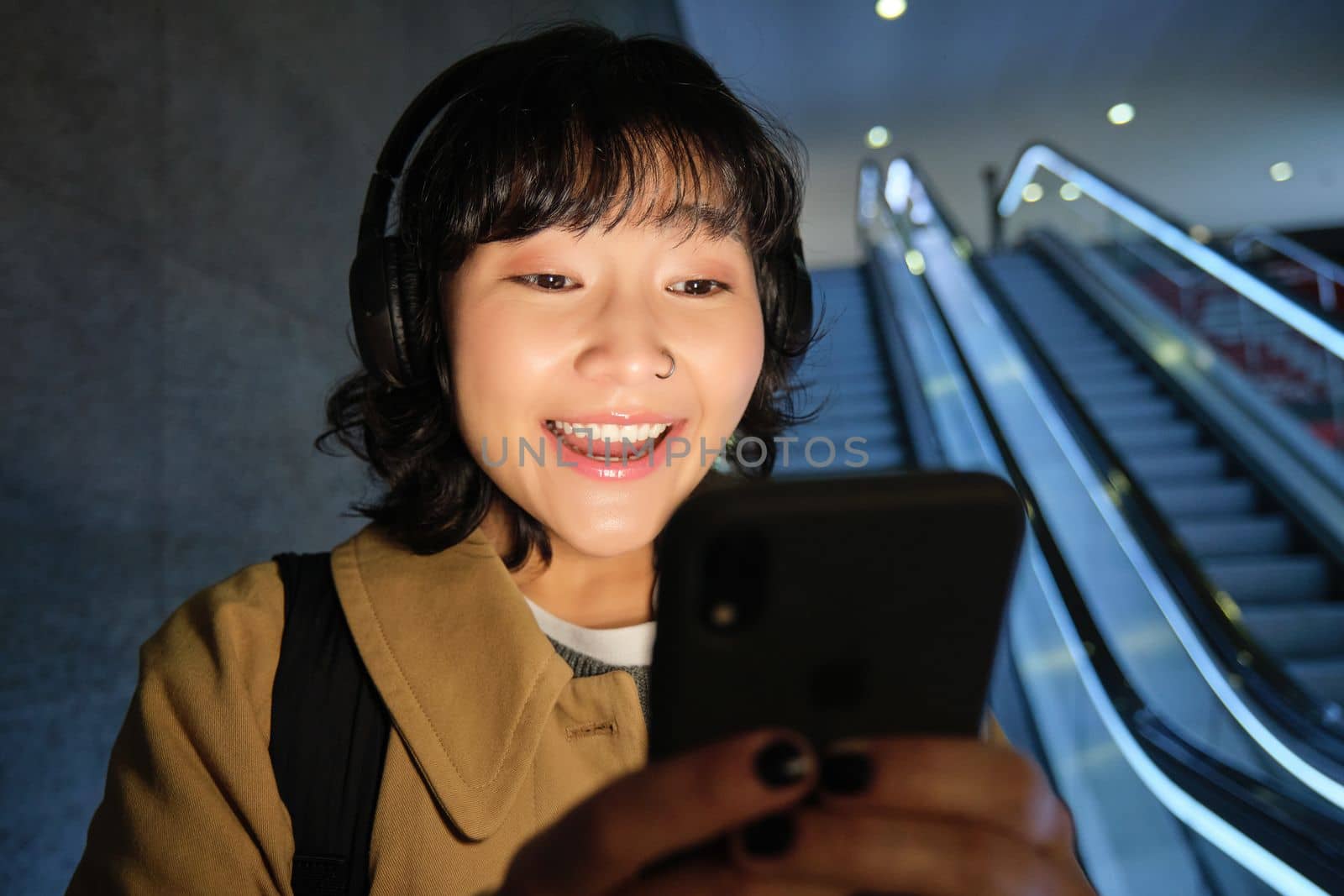 Close up of happy young asian girl, looks at her smartphone with surprised, excited face expression, reading good news on phone, standing on escalator in city.