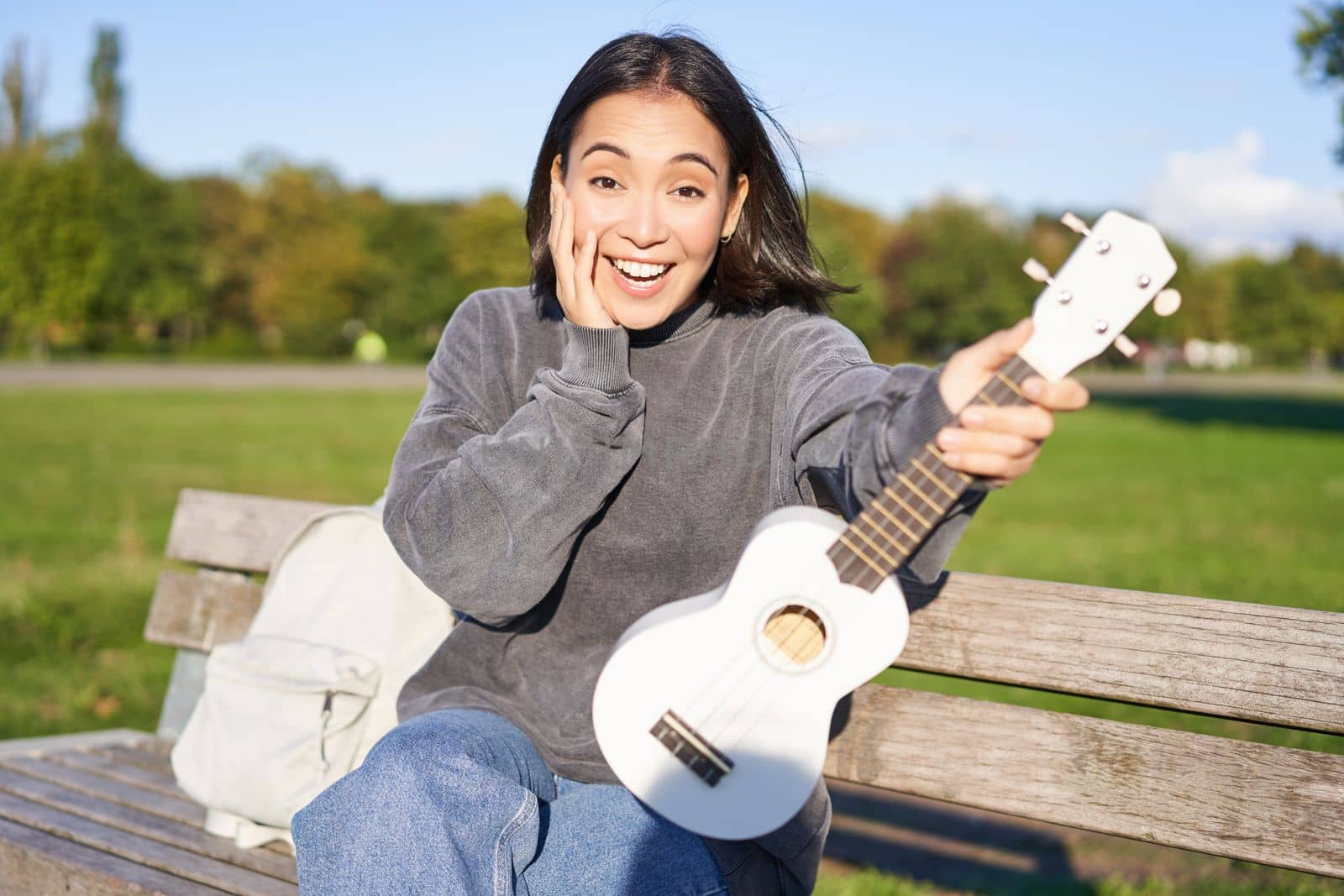 Portrait of young girl musician, sitting in park with ukulele guitar, looking surprised at camera, saying wow.
