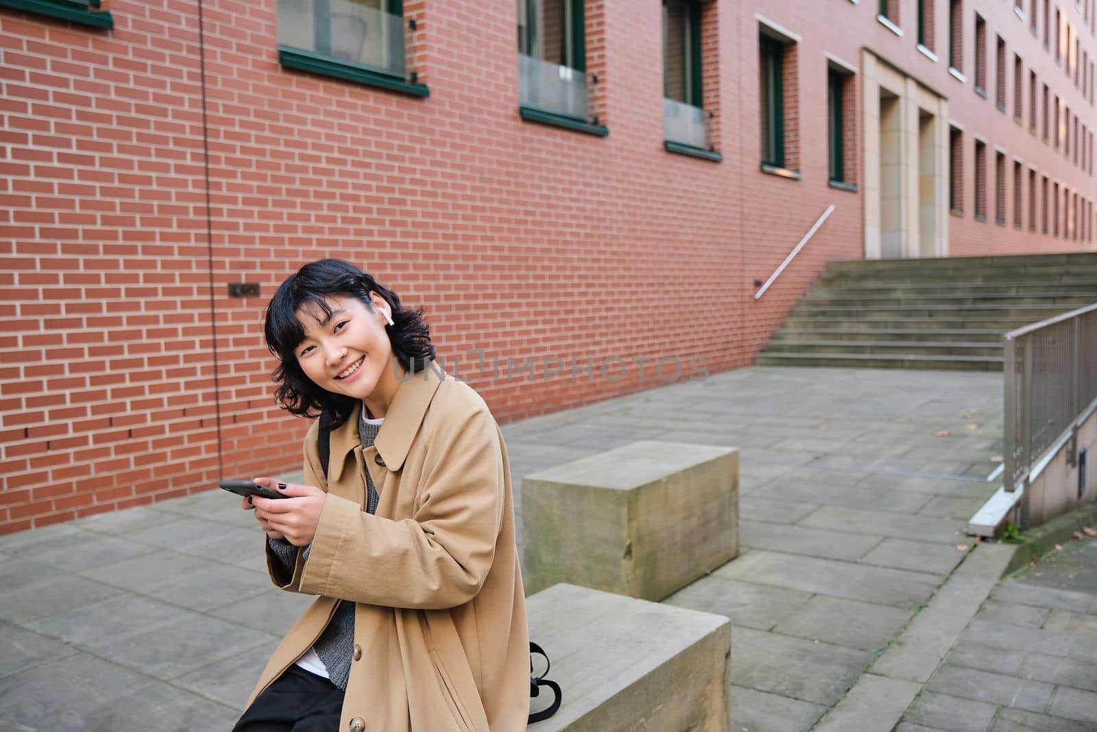 Young asian woman listens music in wireless earphones, uses smartphone on street, walks around city on sunny day, smiling happily.