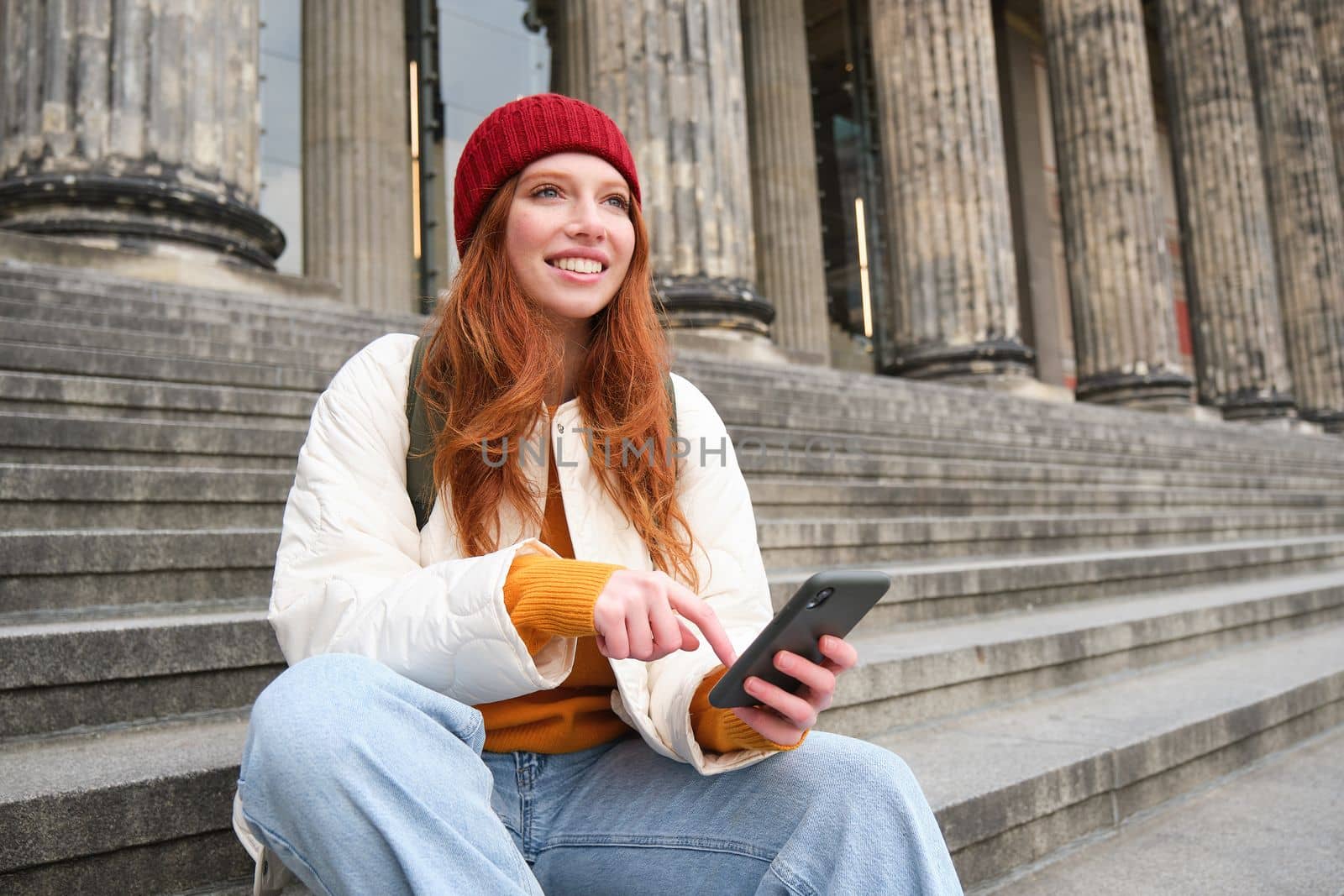 Portrait of young urban girl in red hat, sits on stairs near museum, holds mobile phone, connects to public wifi and surfs net, uses smartphone apps.