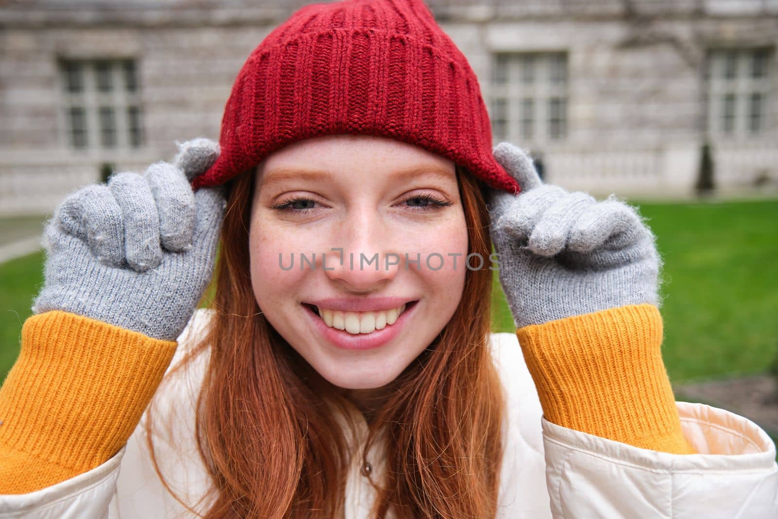 Close up portrait of beautiful redhead woman in red knitted hat, warm gloves, smiling and looking happy at camera, sitting in park.