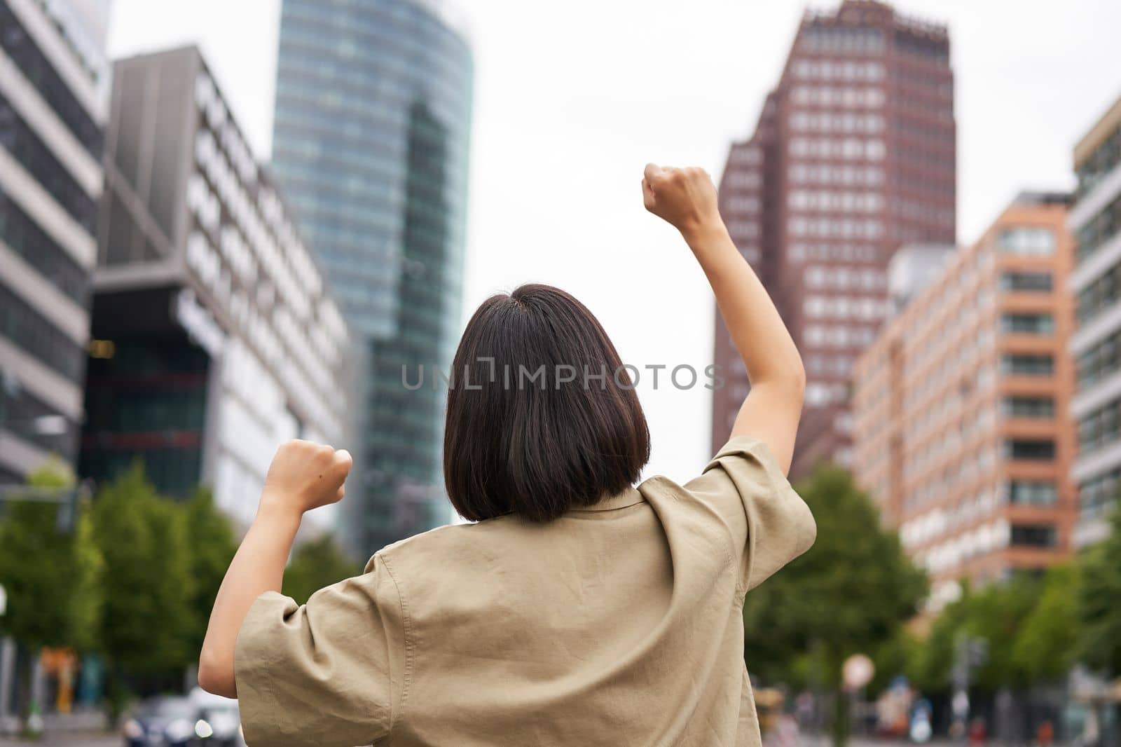 Excitement. Happy girl rear view, raising her hands up and looking at big city skyscrappers, feeling confident, cheering and triumphing by Benzoix
