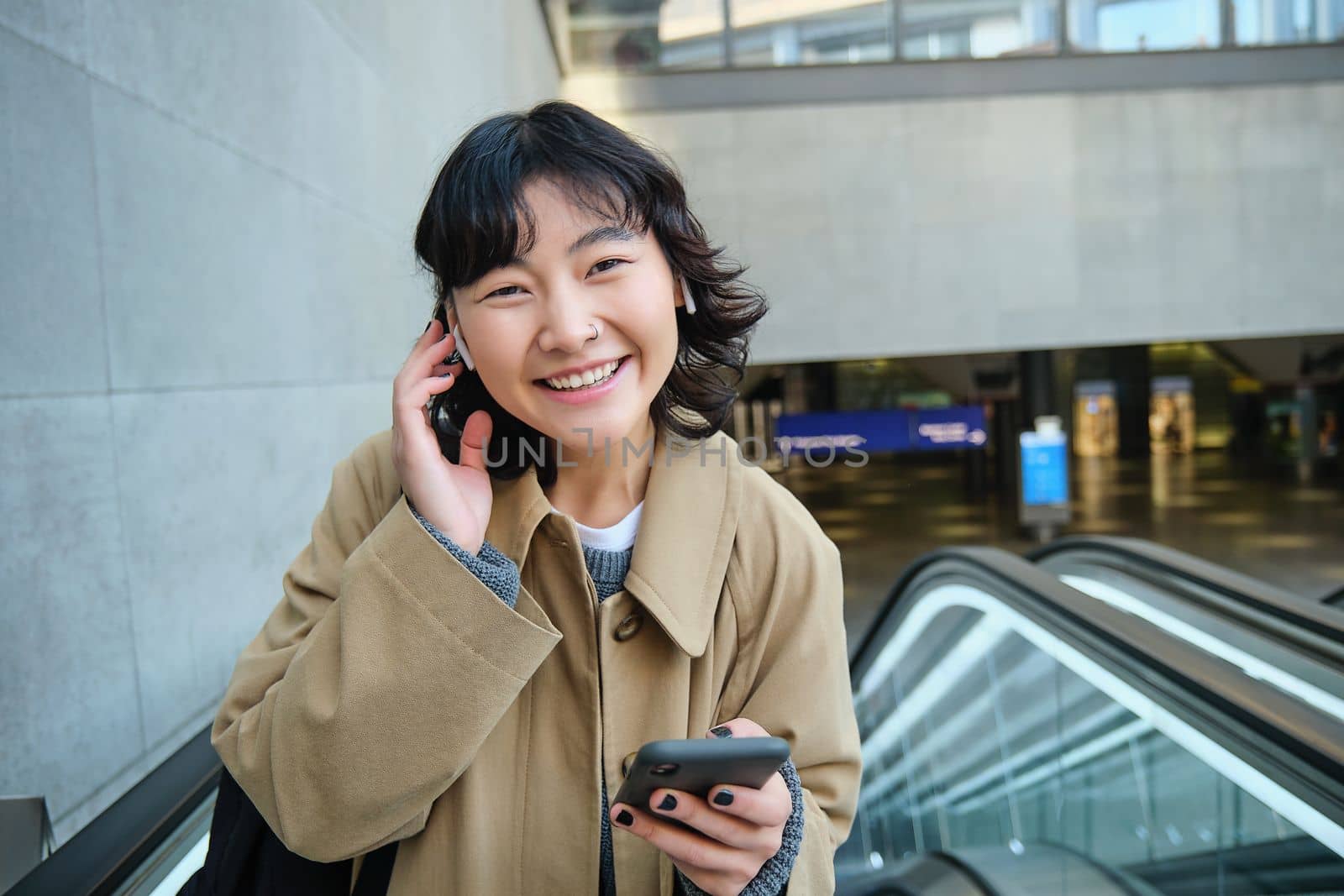 Portrait of cute smiling korean girl, goes up escalator, listens music in wireless earphones and uses mobile phone, holds smartphone.