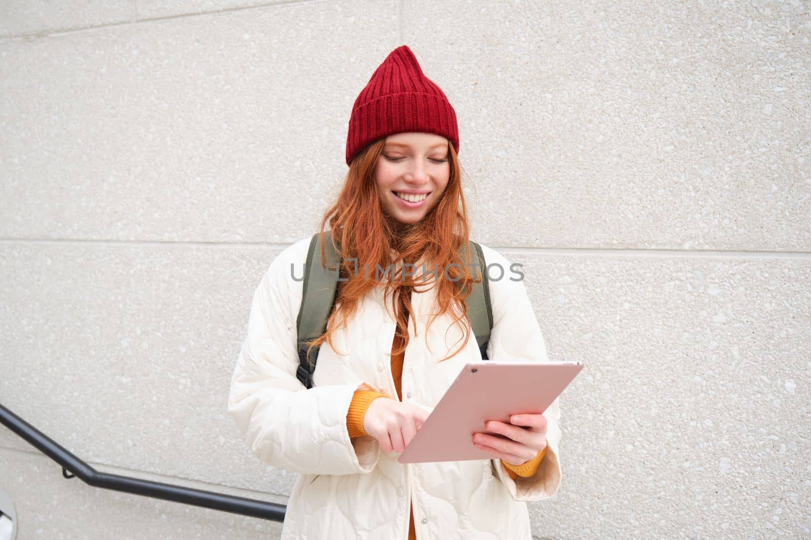 Young redhead woman with red hat, uses her digital tablet outdoors, stands on street with gadget, connects to wifi internet and searches for a location in internet by Benzoix