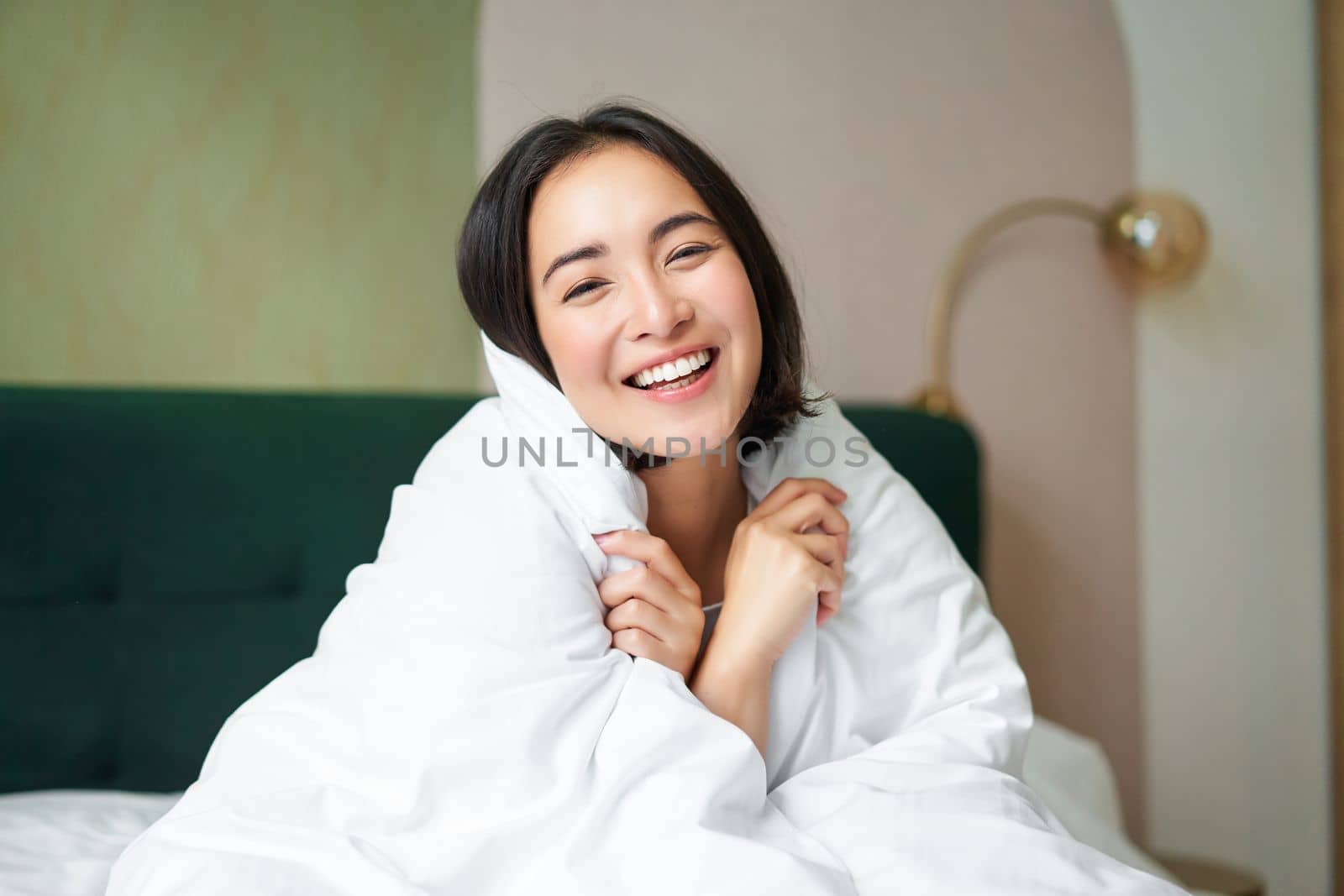 Beautiful romantic woman covers herself with warm duvet, enjoys sleeping and staying in bed, waking up happy in morning, sitting in bedroom.