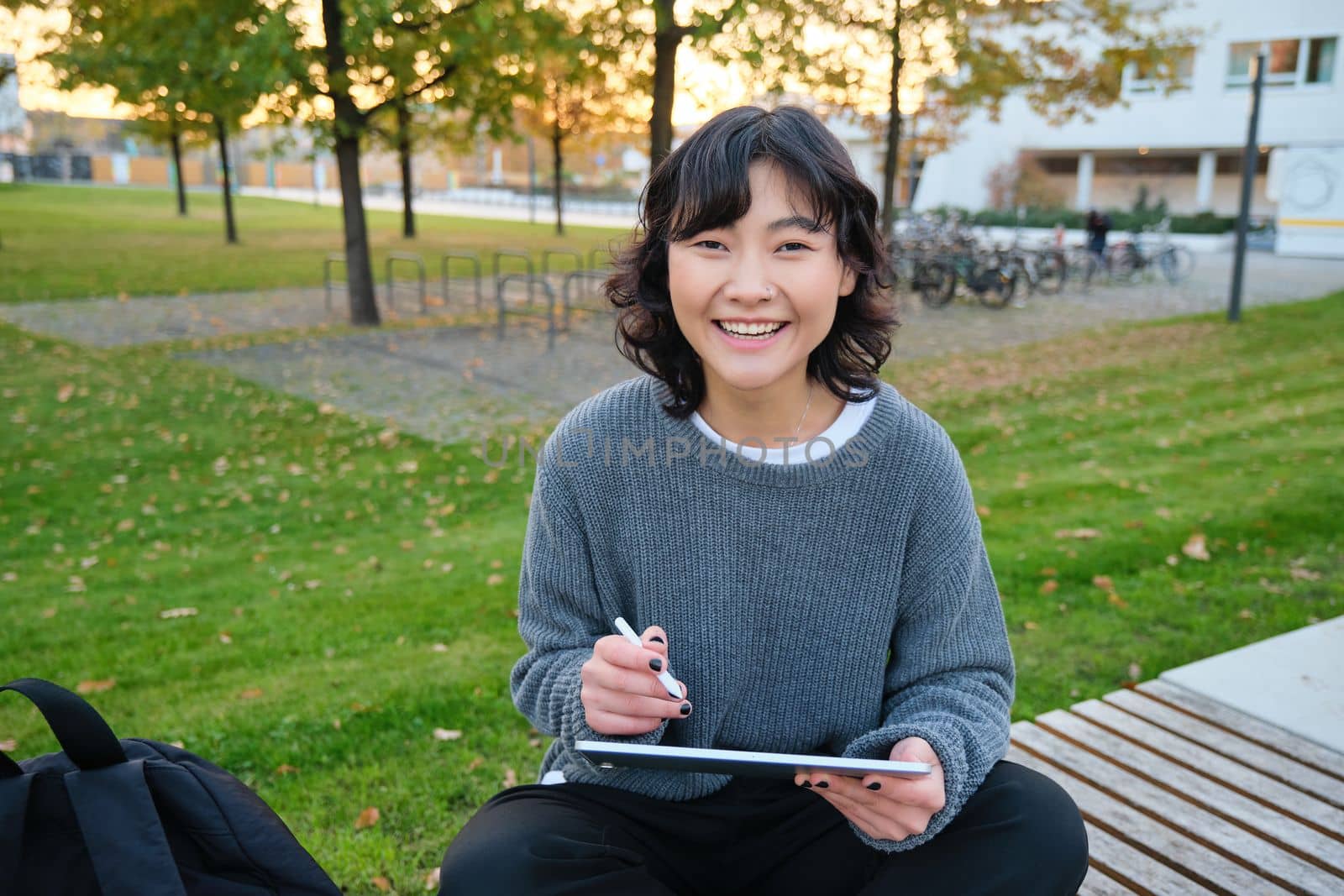 Young girl, korean artist or art student sits in park with digital tablet, draws with graphic pen, scatches a design or project, looks around by Benzoix