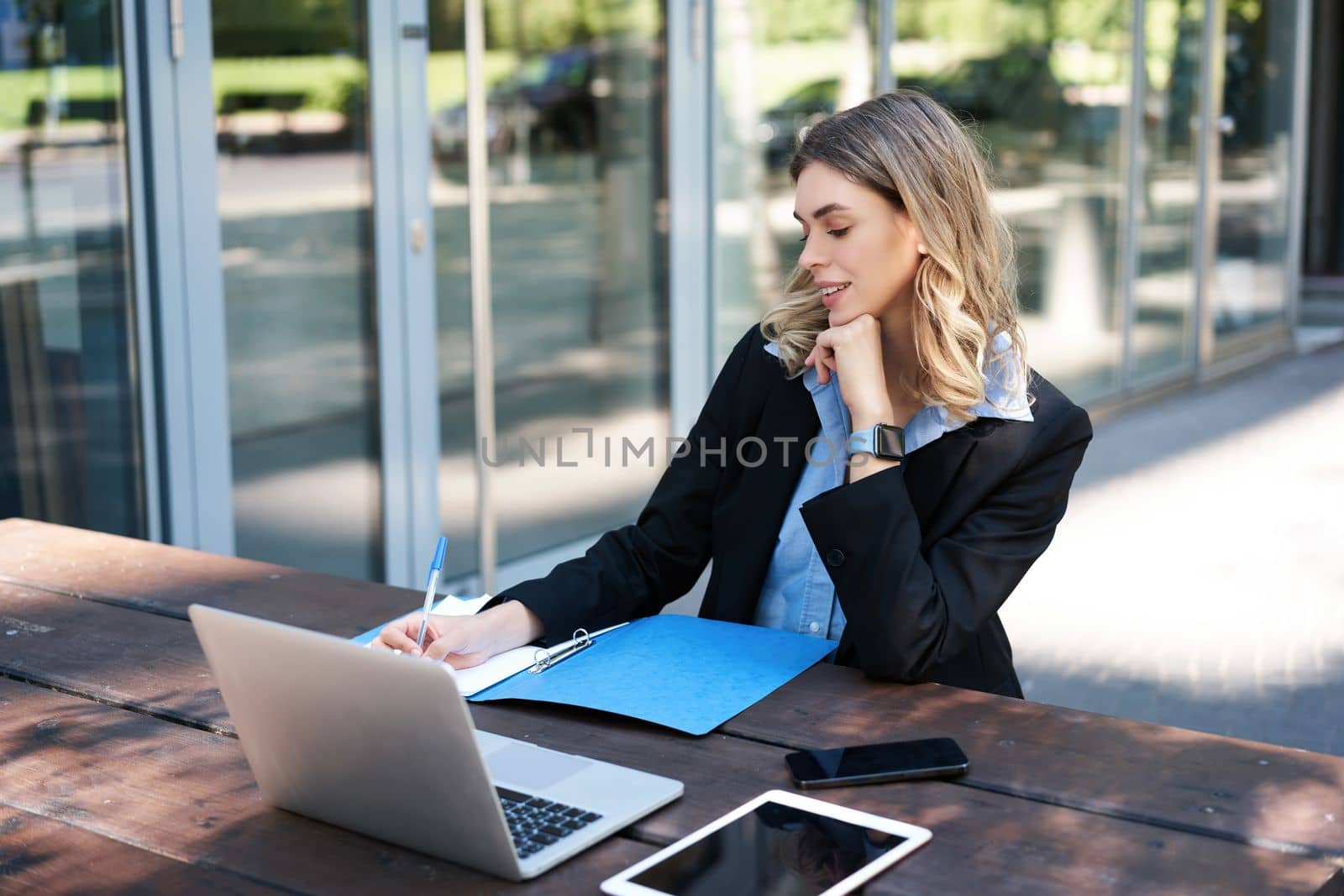 Successful businesswoman working outdoors. Corporate woman sitting on bench with laptop, writing, taking notes during work meeting by Benzoix