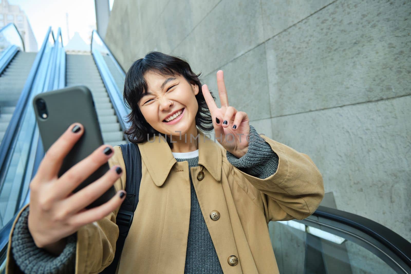 Happy triumphing asian girl, looks amazed and joyful, holds smartphone, goes down escalator with pleased face, wins, hears good news.