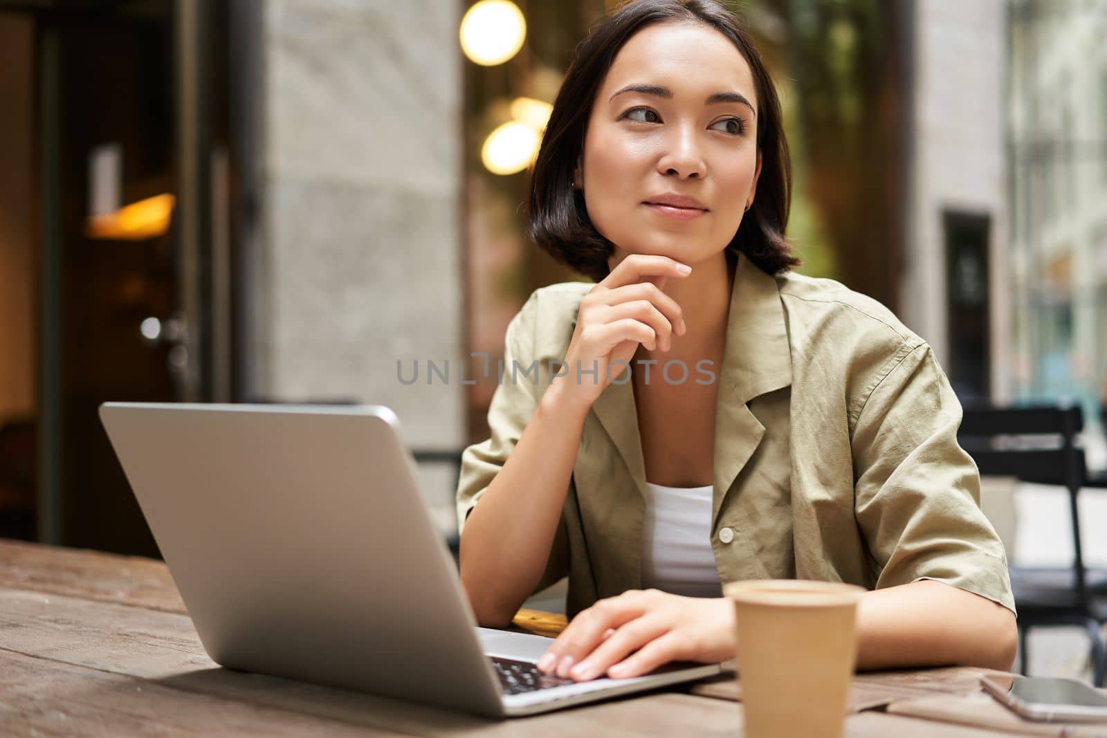 Young woman working in a cafe, using laptop and drinking coffee. Asian girl student with computer studying remotely, sitting on bench near shop.