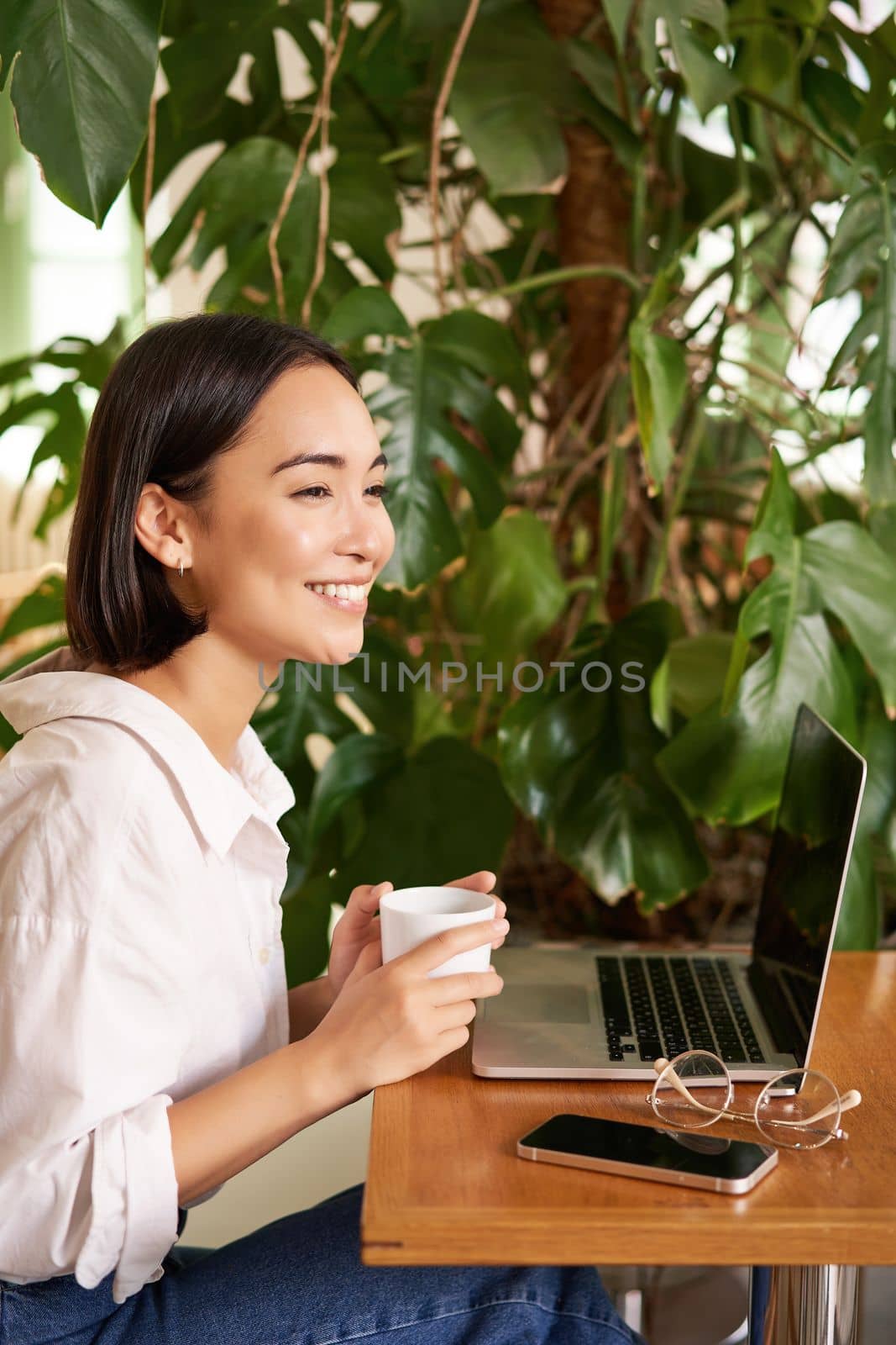 Vertical shot of beautiful woman sits in cafe with laptop, works or studies online at co-working space, smiling relaxed.