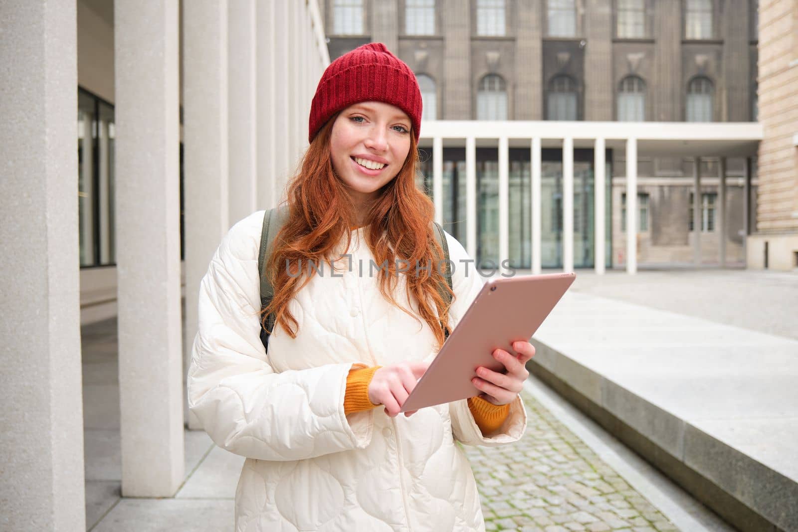 Happy redhead girl in red hat, walks around city with digital tablet, connects to public internet wifi and looks for route, looks at map on her gadget.