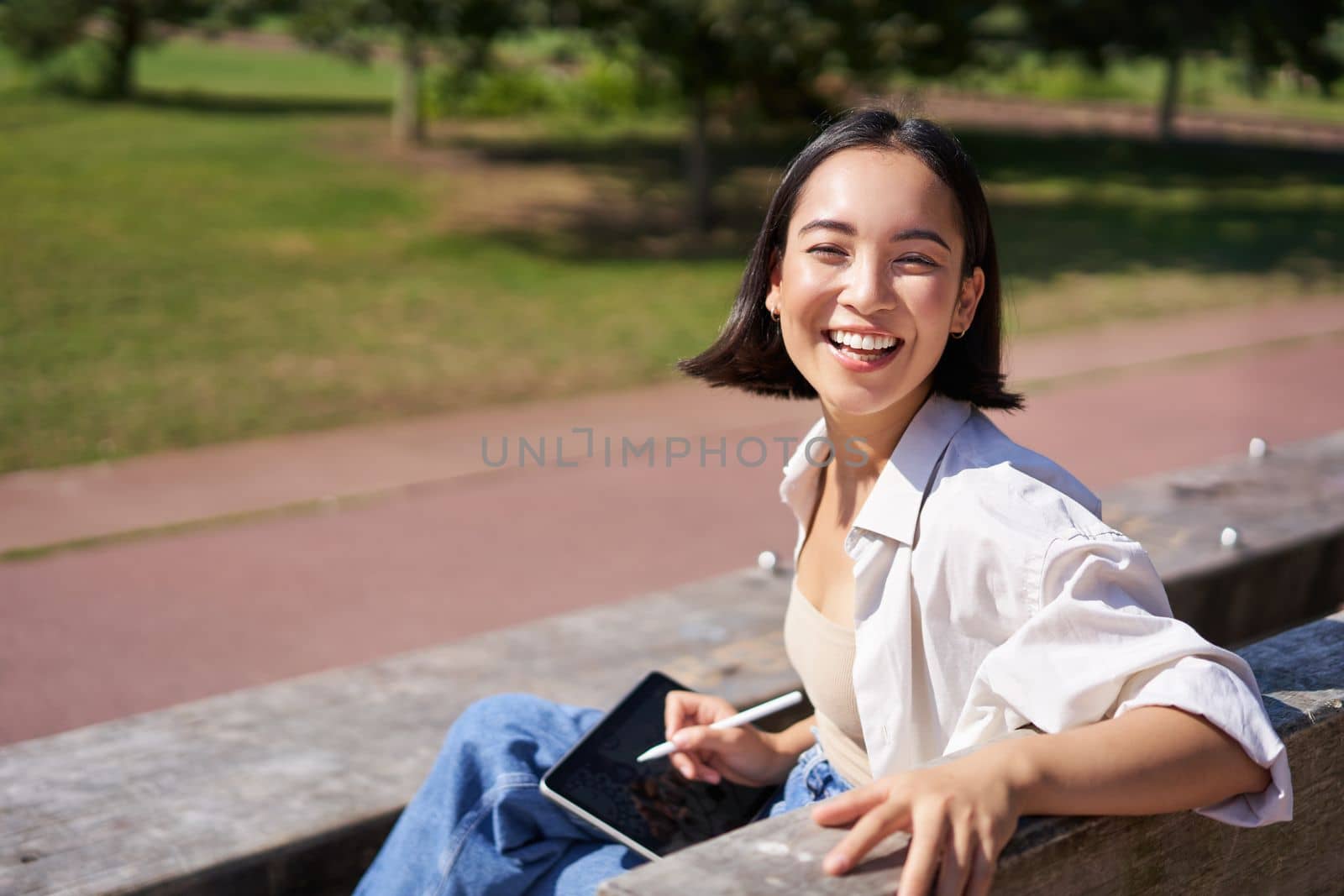 Happy korean woman sitting on bench with digital tablet and graphic pen, smiling, turning back at camera and talking lively.