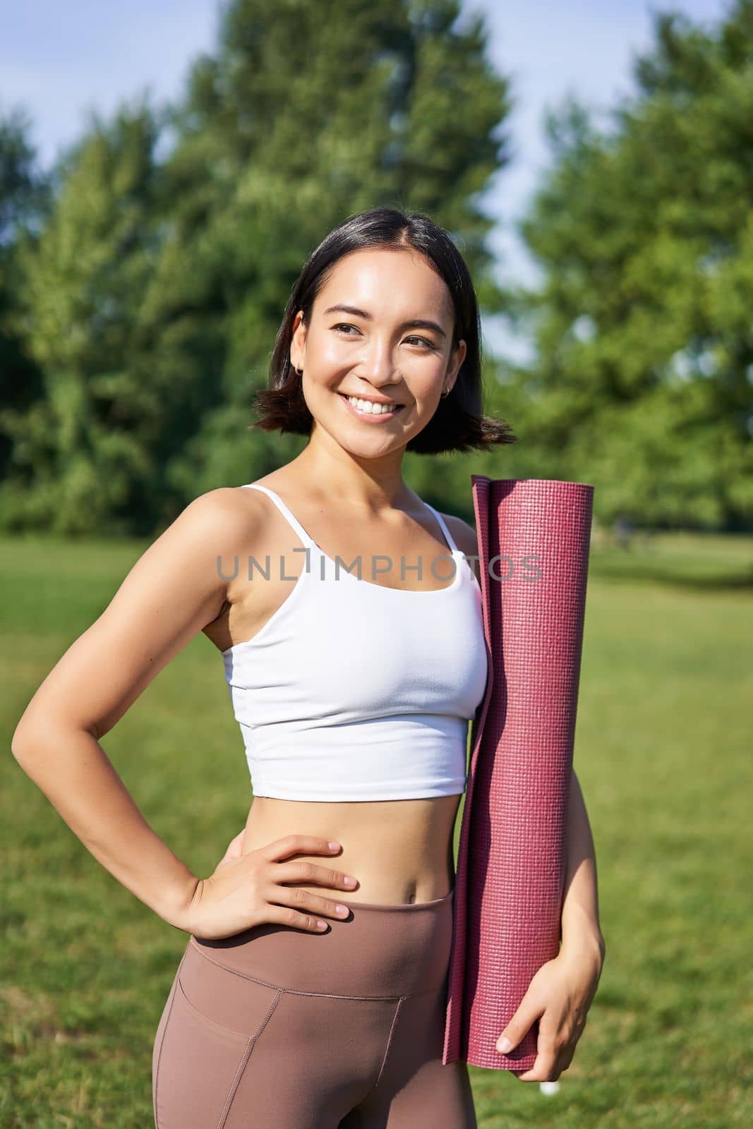 Vertical portrait of young asian fitness girl walks with rubber mat for yoga, goes on training session on fresh air in park, wears sports clothing.