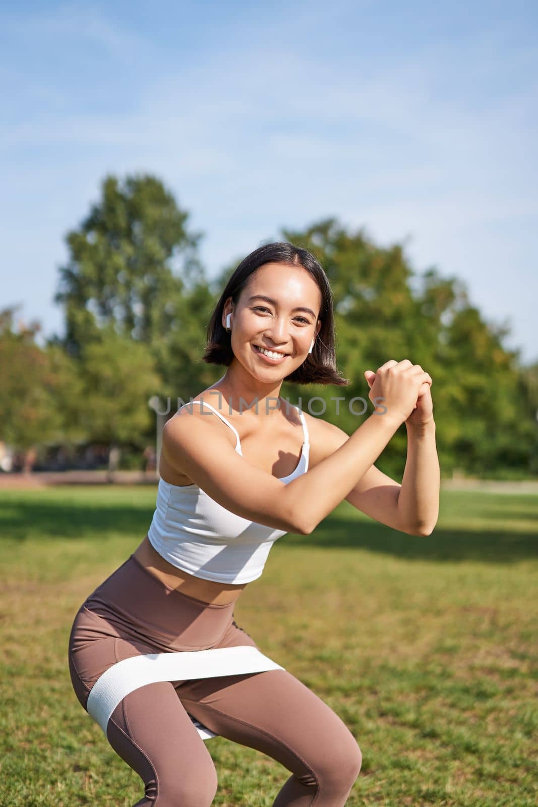 Vertical shot of young fit woman does squats in park, using stretching band on legs, smiling pleased while workout.