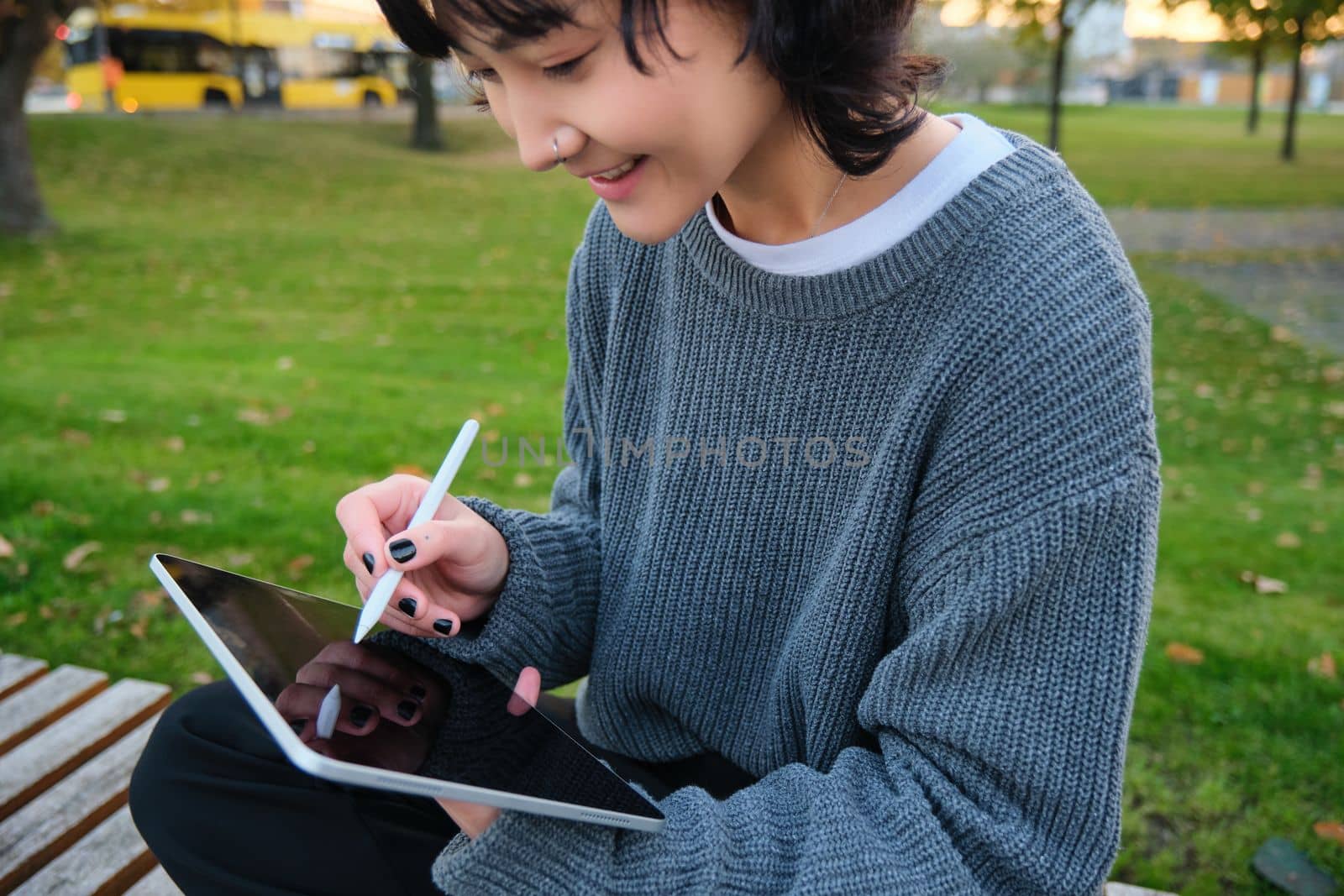 Portrait of young smiling korean girl, graphic designer, artist drawing on digital tablet with a pen tool, sitting in park on fresh air and scatching, taking notes by Benzoix