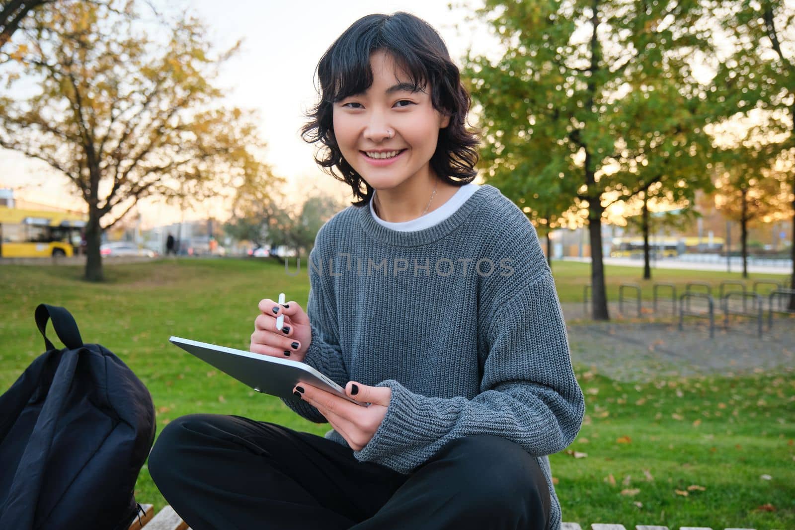 Portrait of young smiling korean girl, graphic designer, artist drawing on digital tablet with a pen tool, sitting in park on fresh air and scatching, taking notes by Benzoix