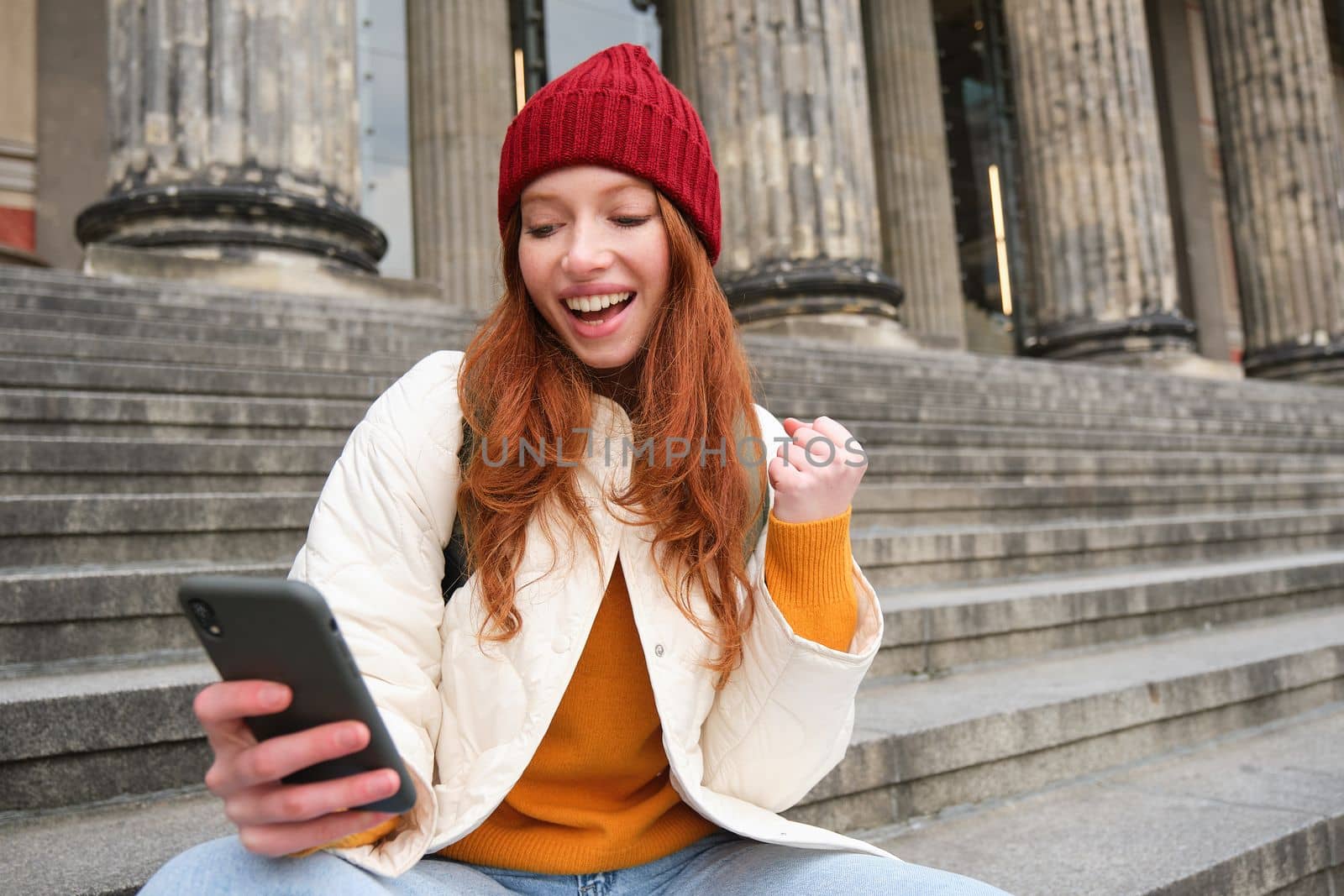 Achievement celebration. Happy redhead girl sits on stairs outdoors and looks at phone, triumphs, wins something and looks satisfied at mobile screen by Benzoix