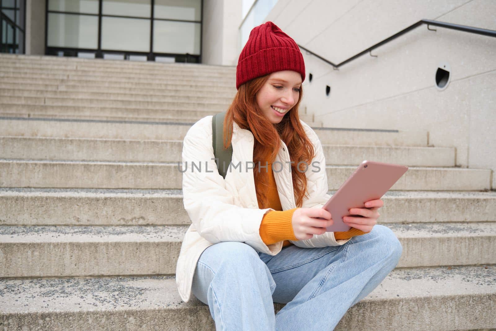 Young stylish girl, redhead female students sits on stairs outdoors with digital tablet, reads, uses social media app on gadget, plays games while waits on street by Benzoix