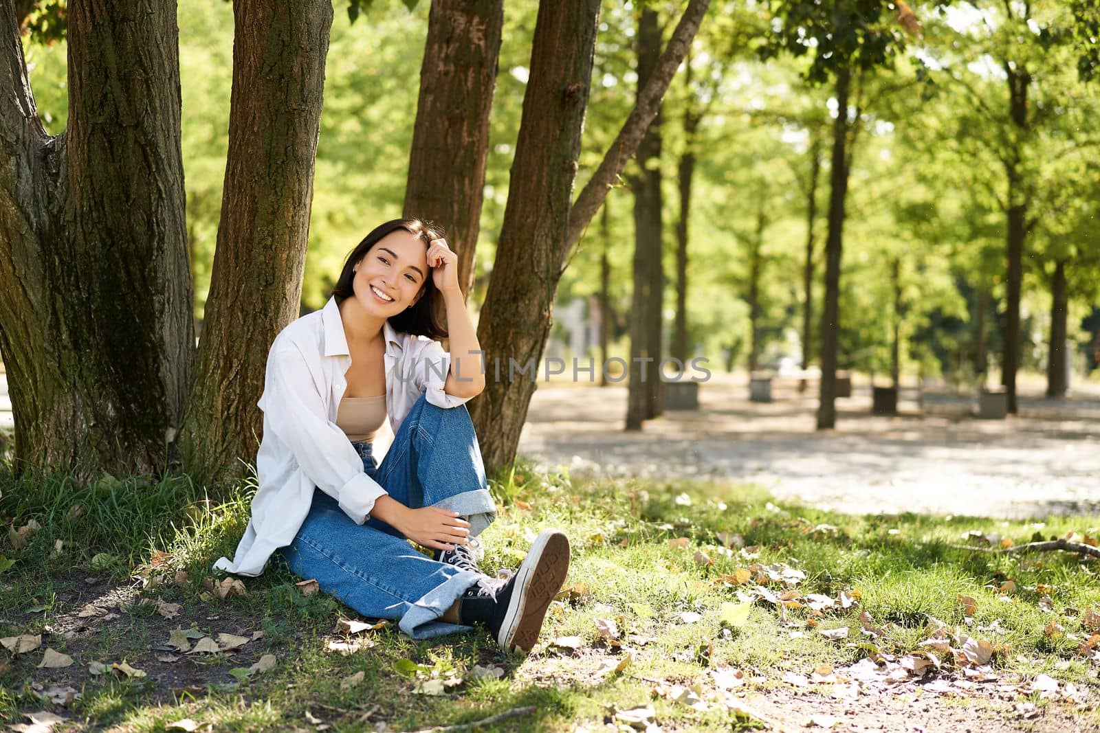 Young people. Beautiful asian girl sits near tree in park and rests, smiling and looking into distance, relaxing outdoors on fresh air in summer by Benzoix