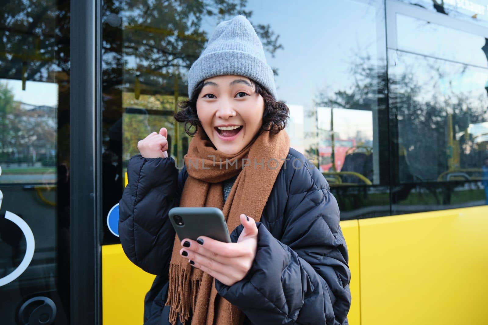 Enthusiastic girl rejoice, reads message on mobile phone and celebrates, stands near her bus on public transport stop and looks excited, posing in warm winter clothes by Benzoix